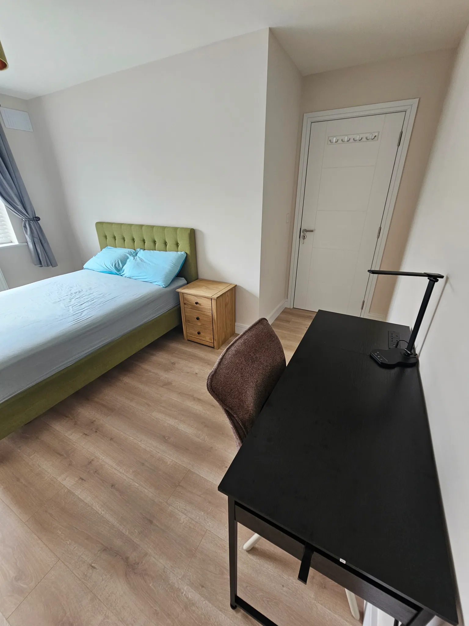 Bedroom with a double bed with a green upholstered headboard, a wooden nightstand, a desk with a chair and desk lamp, window with curtains, and a closed door.