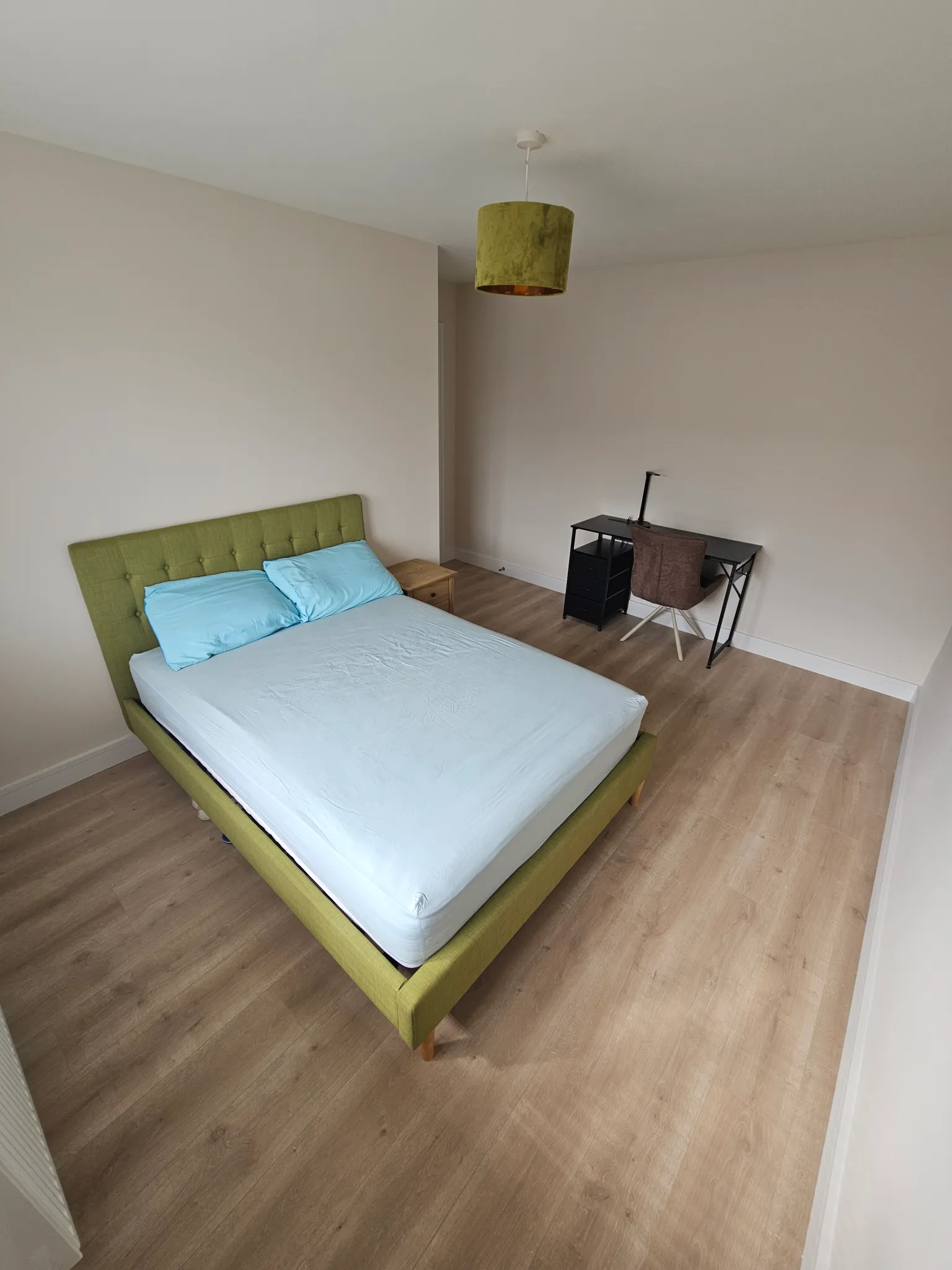 Bedroom with a double bed with a green upholstered headboard, a bedside table, a desk with a chair and lamp, and a ceiling pendant light.