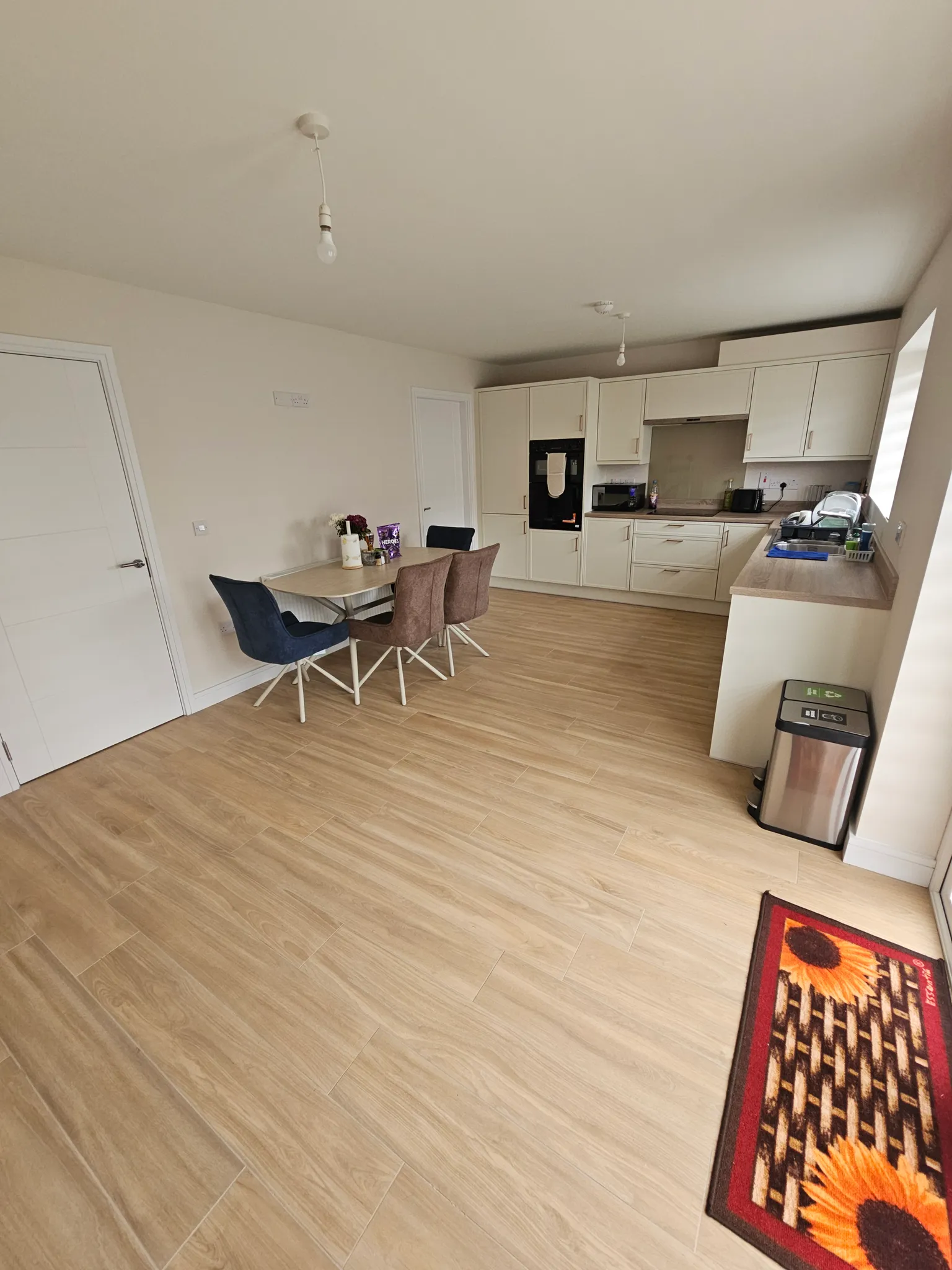 Open-plan kitchen with light wood floors, white cabinets, built-in oven, countertop with sink and appliances, and a small dining table with four chairs.