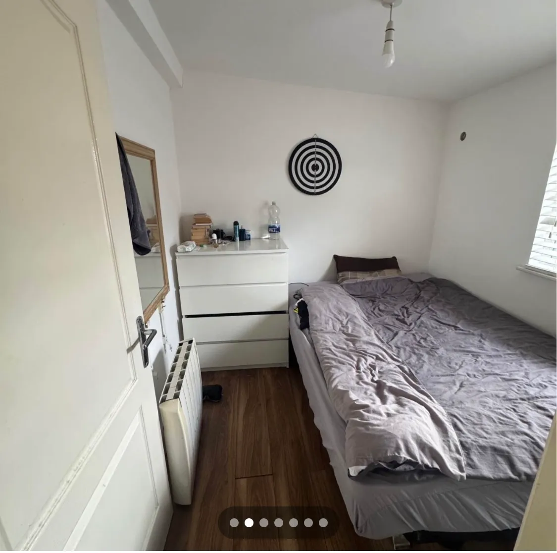 Bedroom with a double bed, gray bedding, white three-drawer dresser, wall target decoration, window with blinds and a hardwood floor.