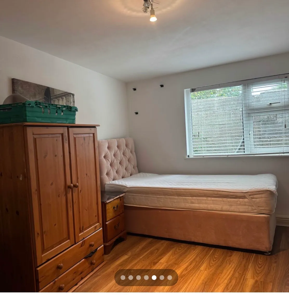 Bedroom with a single bed with tufted headboard, wooden wardrobe, small bedside drawers, and a window with blinds.