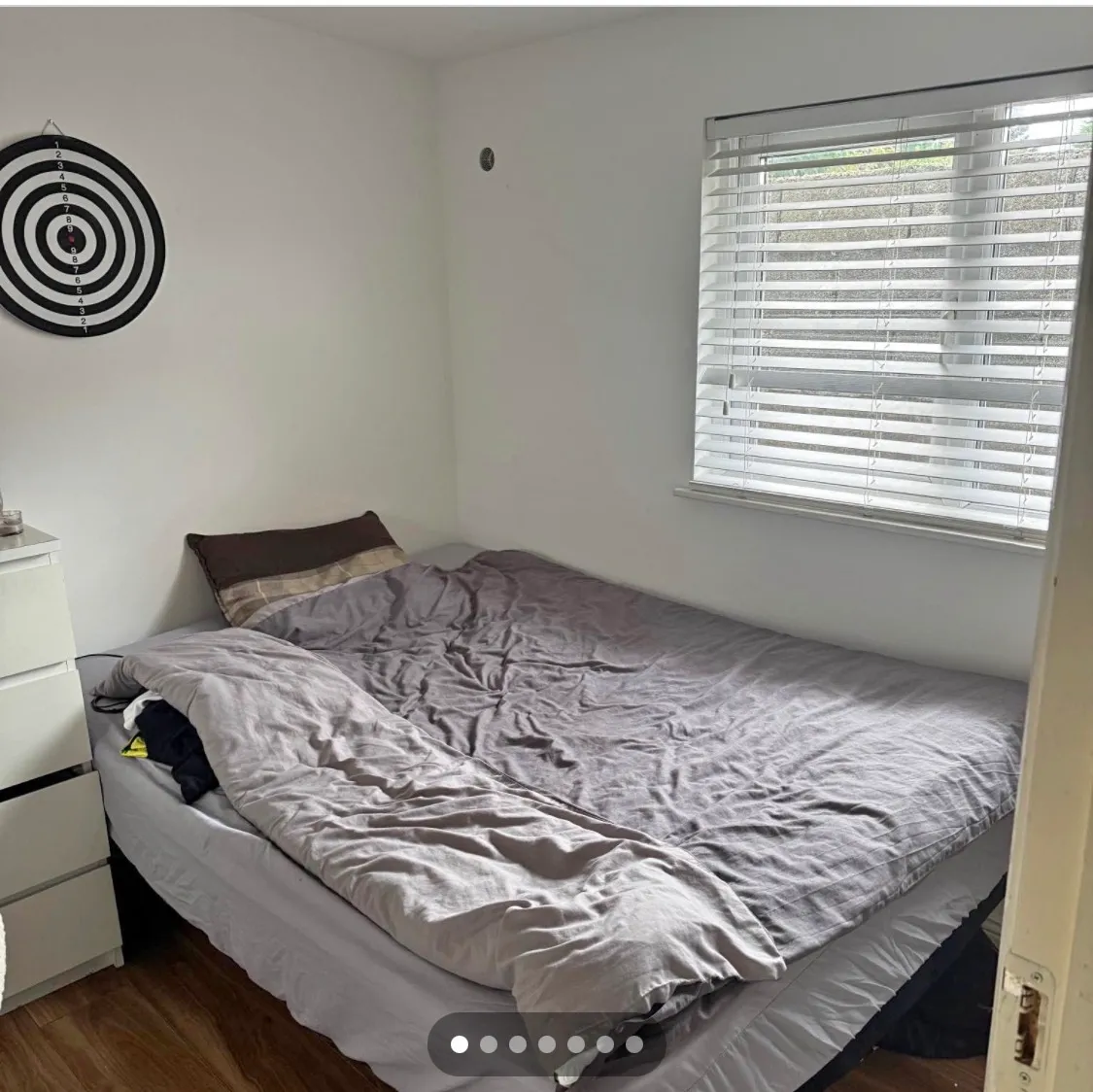 Bedroom with a double bed, gray duvet, pillow, window with horizontal blinds, white three-drawer dresser, and a circular target on the wall.