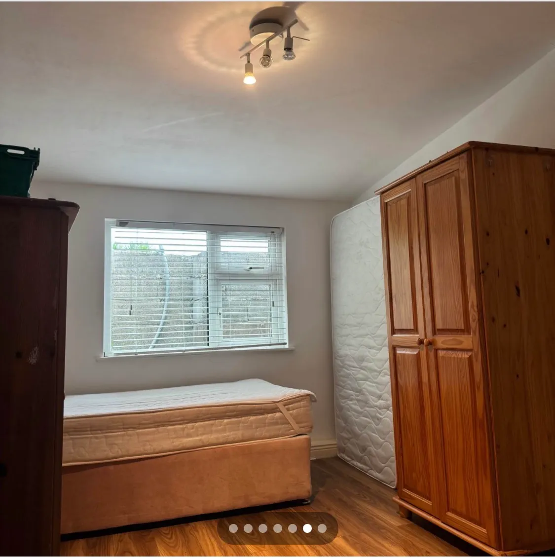 Bedroom with a single bed, window with blinds, upright spare mattress, and wooden wardrobe on a wood floor.