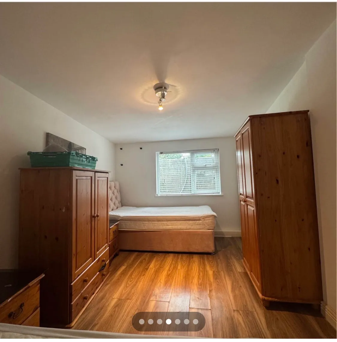 Bedroom with a single bed and mattress against a wall, two wooden wardrobes and a chest of drawers, window with blinds, and ceiling light.