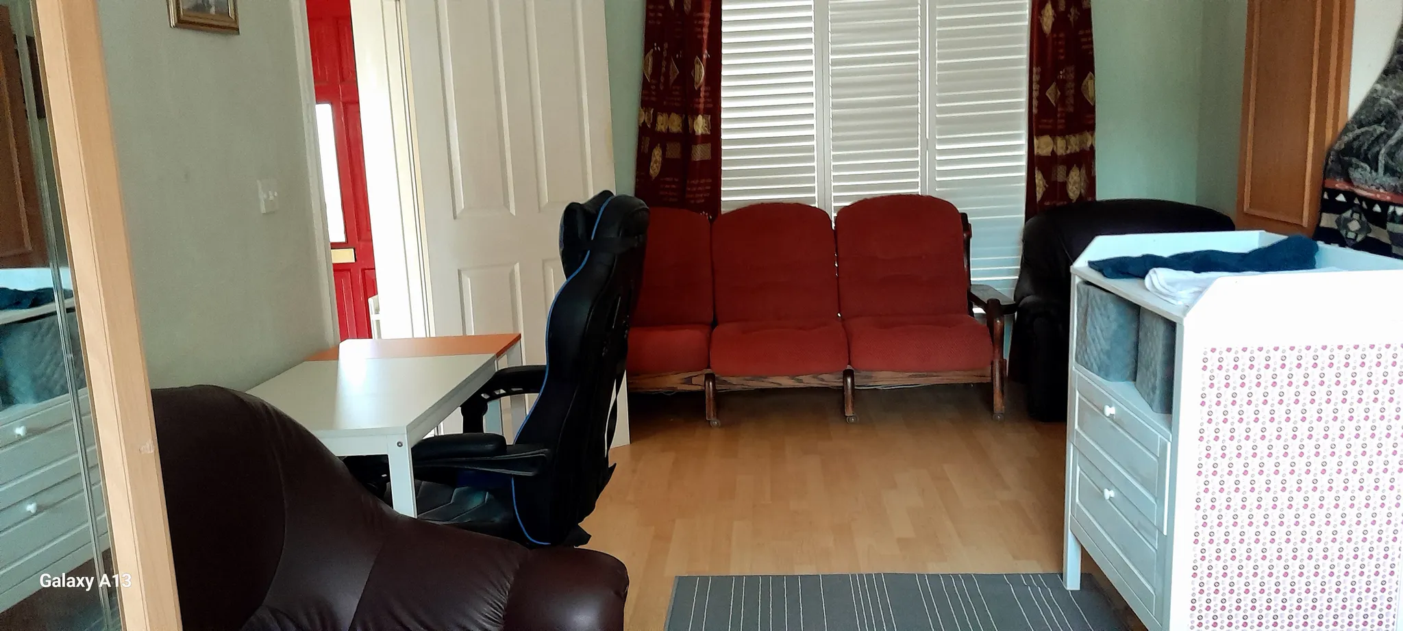 Living room with three red connected seats, a black office chair at a white desk, brown armchairs, white shuttered window and a white dresser at right