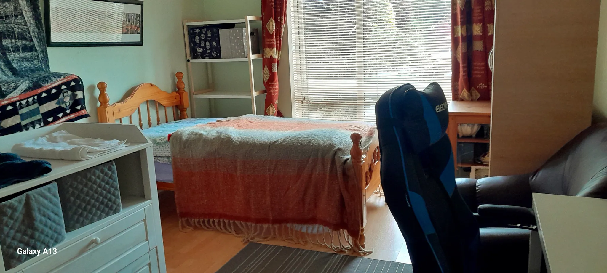 Bedroom with a single wooden bed covered by a blanket, window with horizontal blinds and curtains, shelving, wardrobe, desk, and two chairs (one gaming-style).