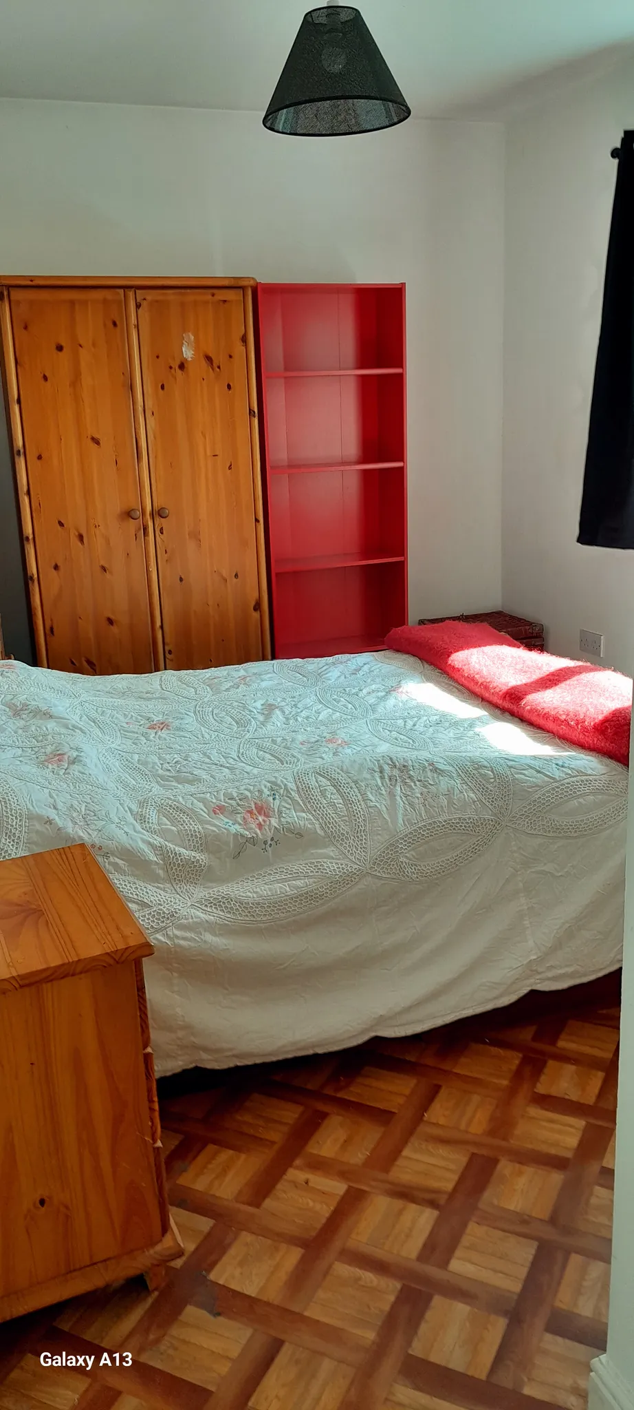 Bedroom with a double bed, wooden wardrobe, red shelving unit, small wooden bedside table, black pendant lamp, and parquet floor; sunlight on the bed and a dark curtain at the window.