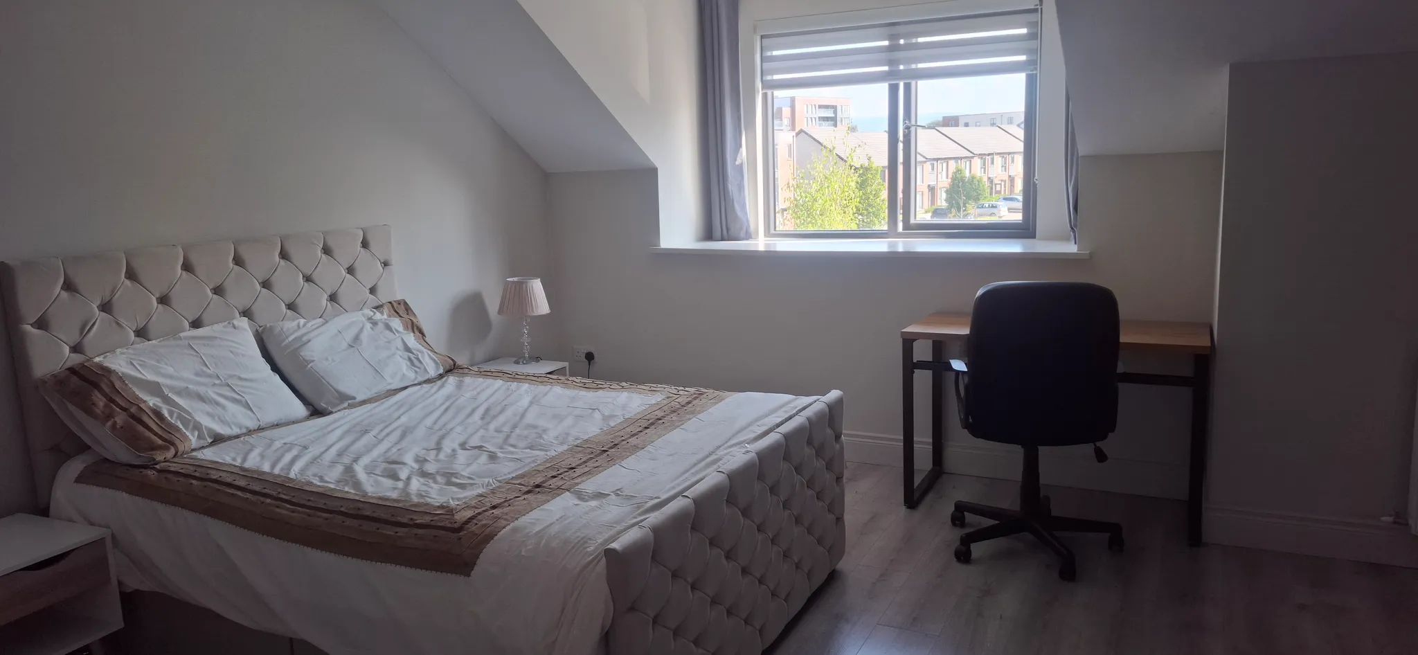 Bedroom with a double bed and tufted headboard, bedside table with lamp, window with blinds and a desk with an office chair