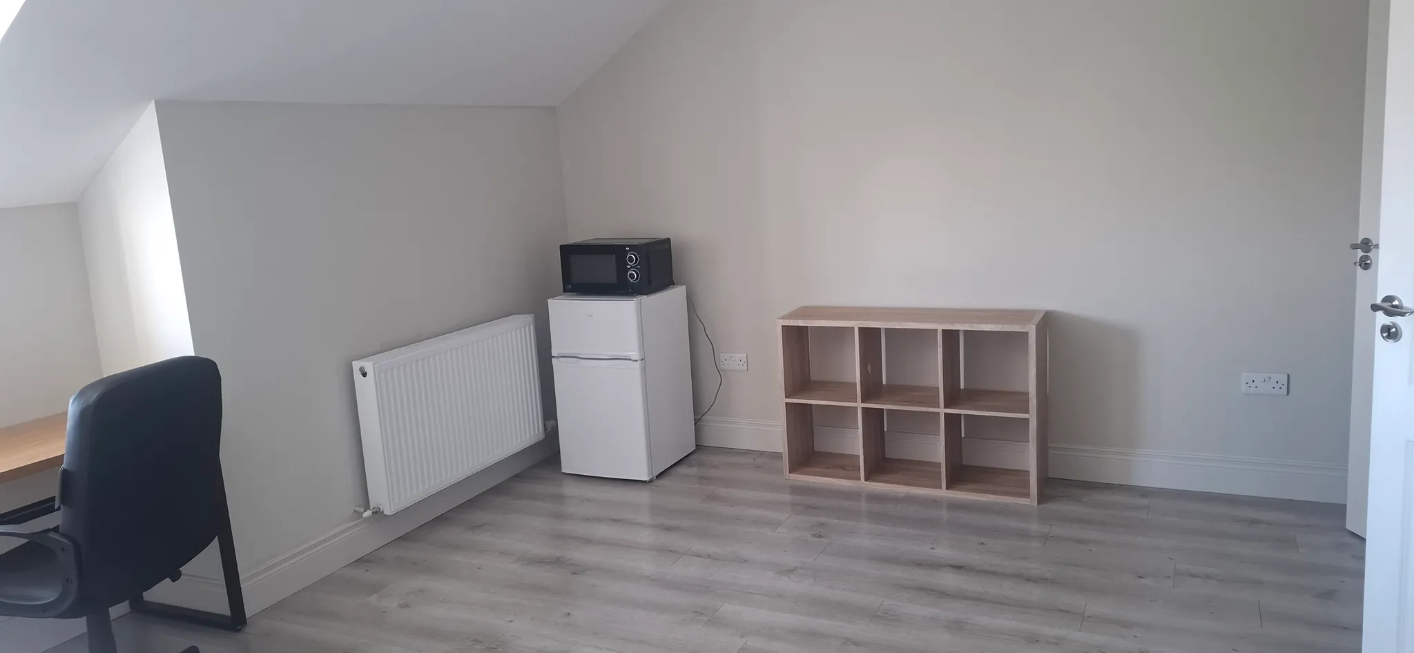Room corner with a small refrigerator topped by a microwave, a six-cube wooden shelf, a desk chair, radiator and visible power outlets on a laminate floor.