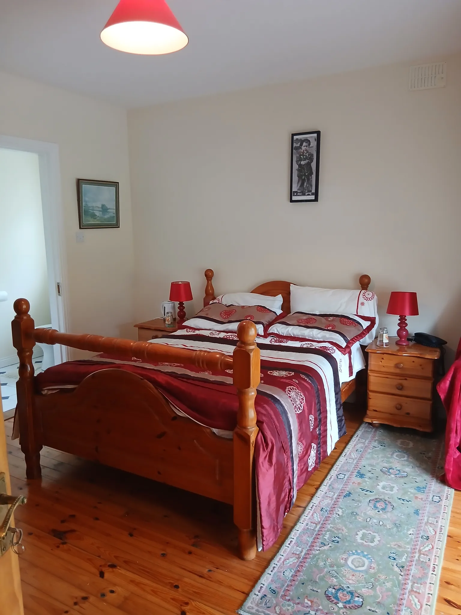 Bedroom with a wooden double bed, patterned duvet, two bedside tables with red lamps, a framed picture on the wall and a long rug on a hardwood floor.