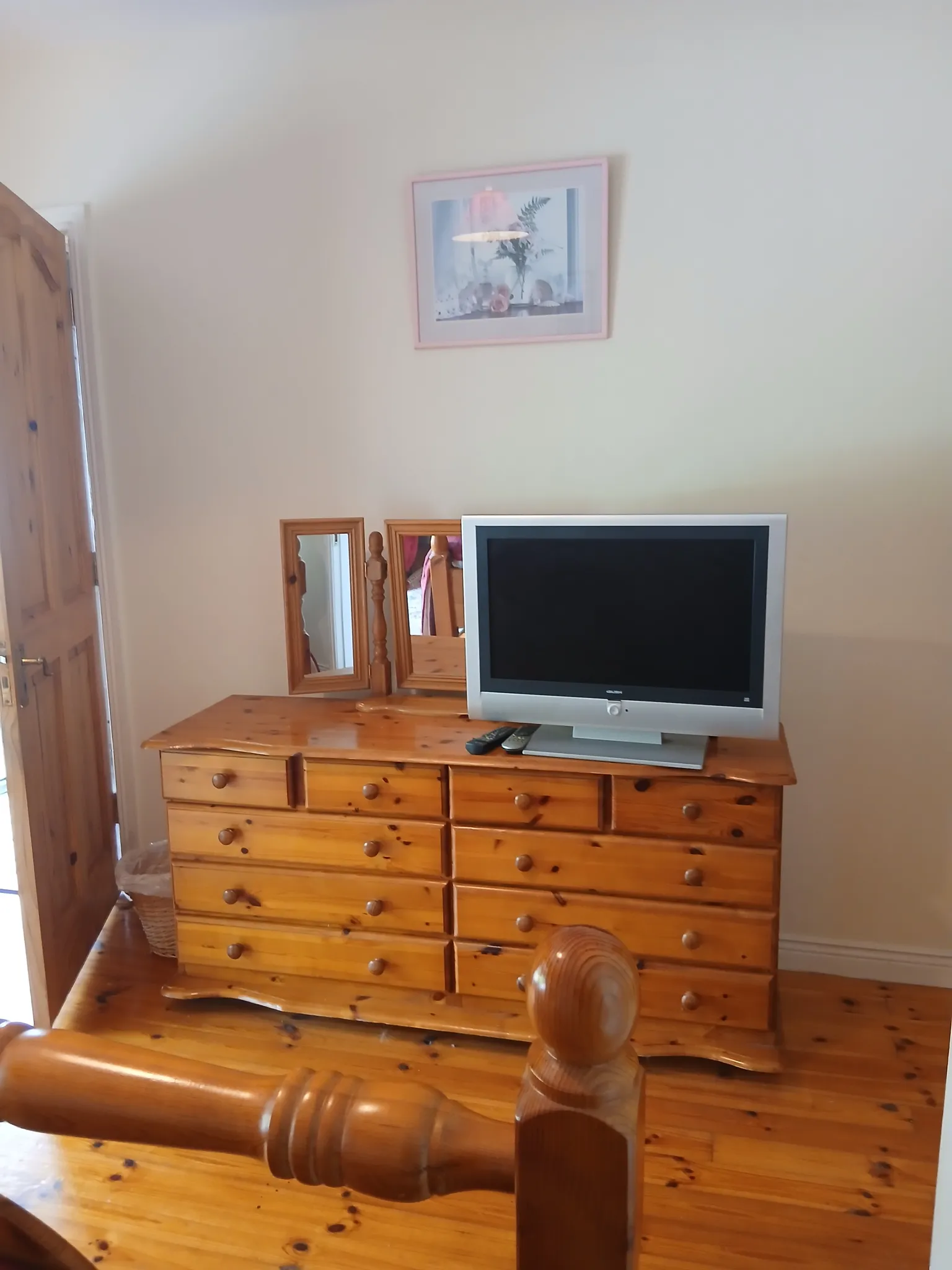 Bedroom showing a wooden dresser with a television, three-panel mirror, framed wall art, and a wooden bedpost in the foreground.