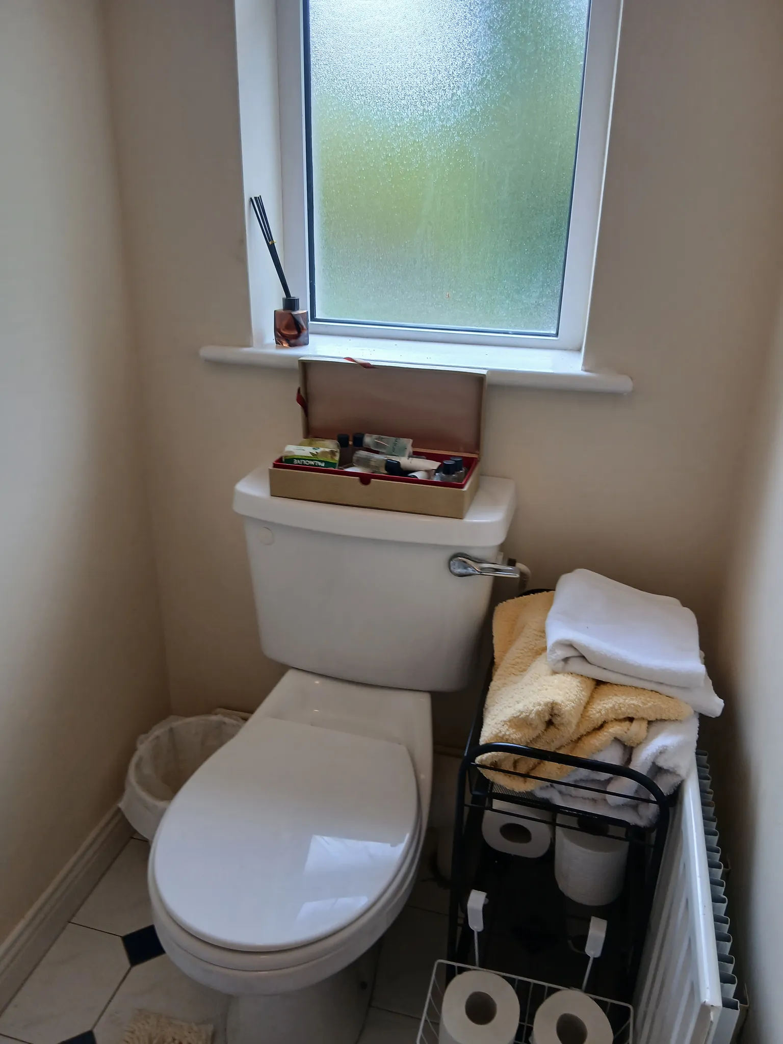 Small bathroom showing a toilet under a frosted window, a box of toiletries on the cistern, and a rack with folded towels and toilet paper.