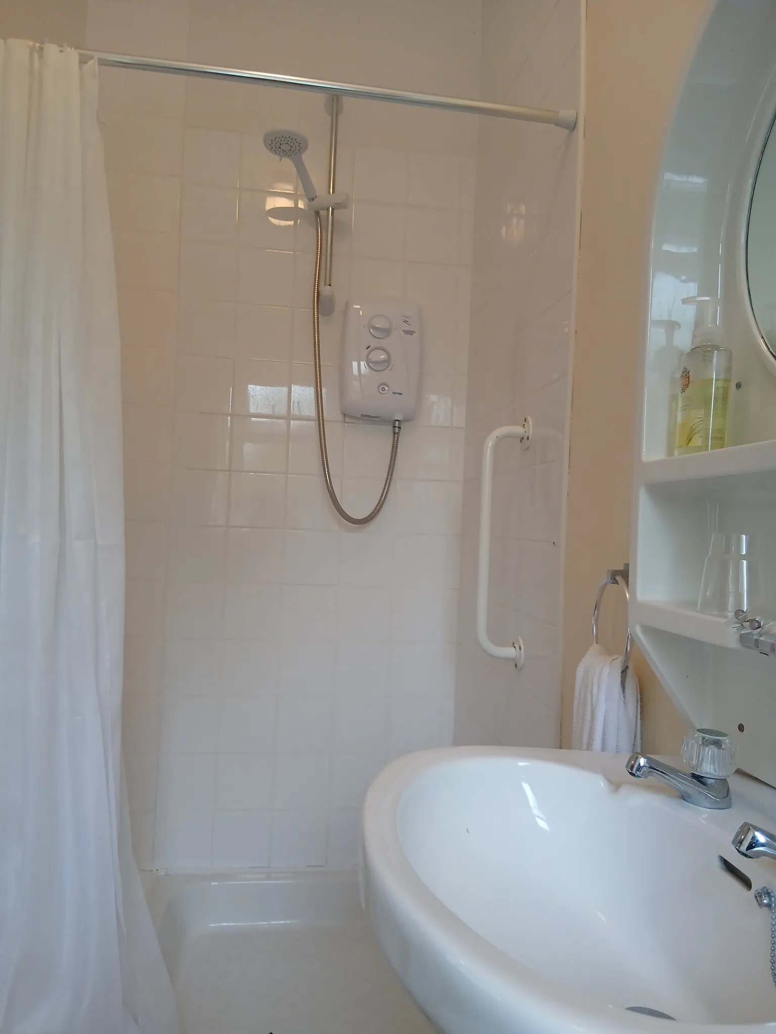 Bathroom with a tiled shower area, handheld showerhead and curtain, wall-mounted shower unit, grab bar, sink with faucet, and shelves with soap dispenser and glass.