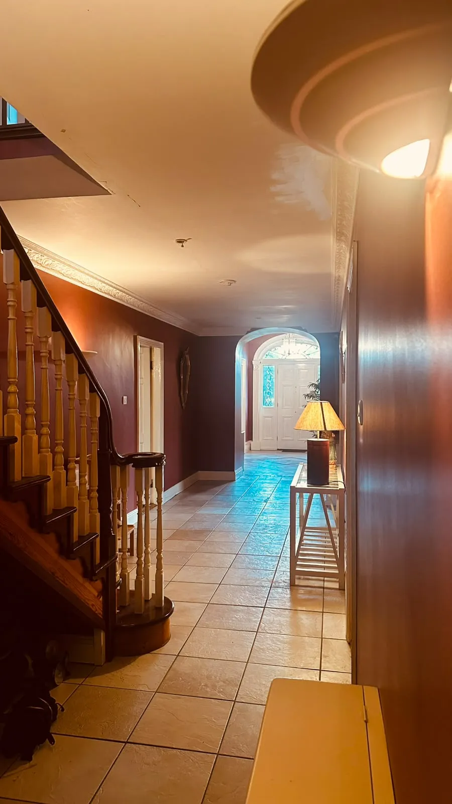 Hallway with a tiled floor, wooden staircase and banister on the left, console table with a table lamp on the right, and a front door visible through an arched doorway at the far end.