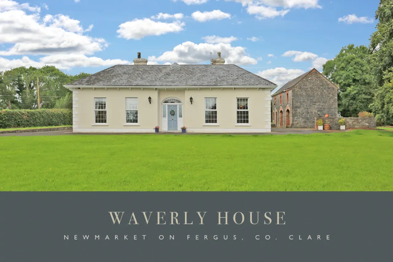 Exterior front facade of a single-story house with a centered front door, three front windows, a large lawn in foreground, and a stone outbuilding to the right under a partly cloudy sky.