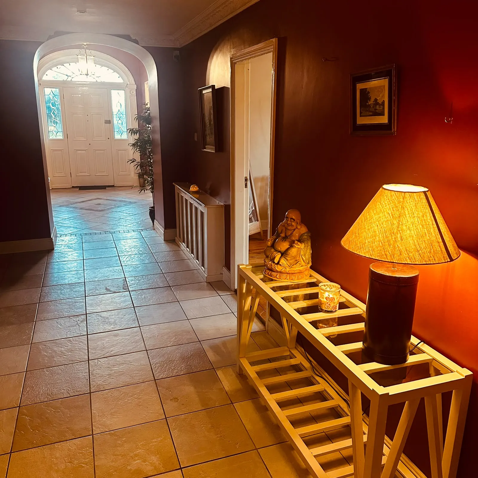 Hallway with tiled floor leading to a white front door with sidelights; wooden console with lamp and decorative statue along a red wall.