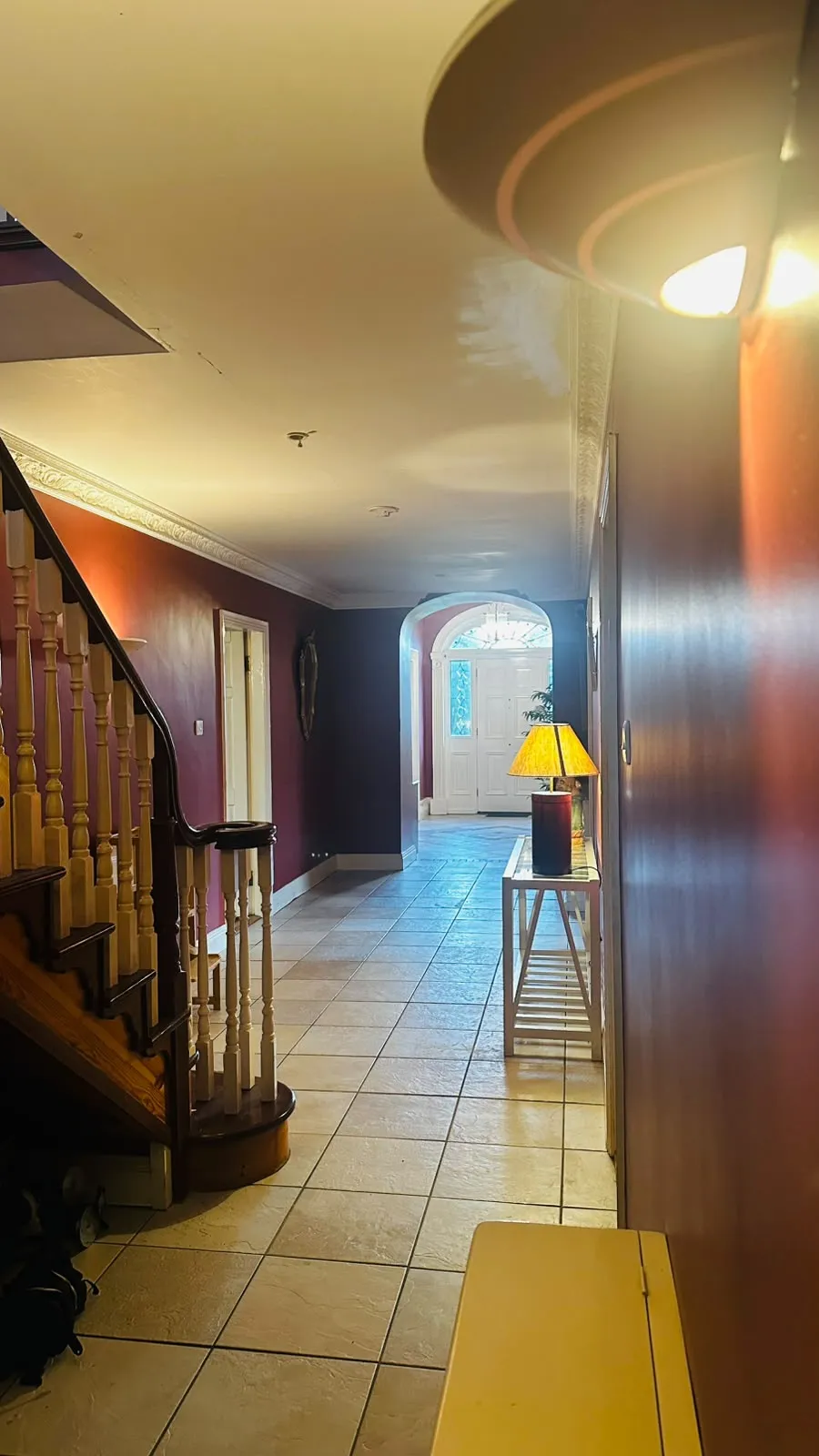 Hallway with tiled floor, wooden staircase on the left, console table with a lamp on the right, and an arched entry leading to a front door.