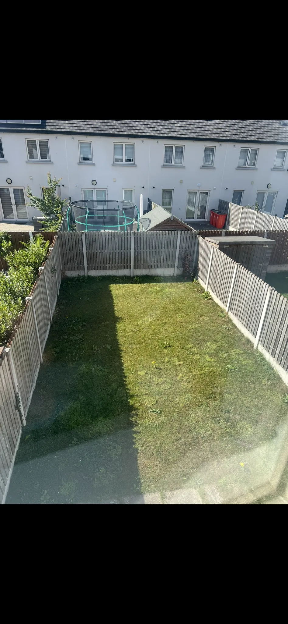Fenced backyard with a grass lawn, wooden shed, trampoline, and neighboring row houses visible beyond the fence.