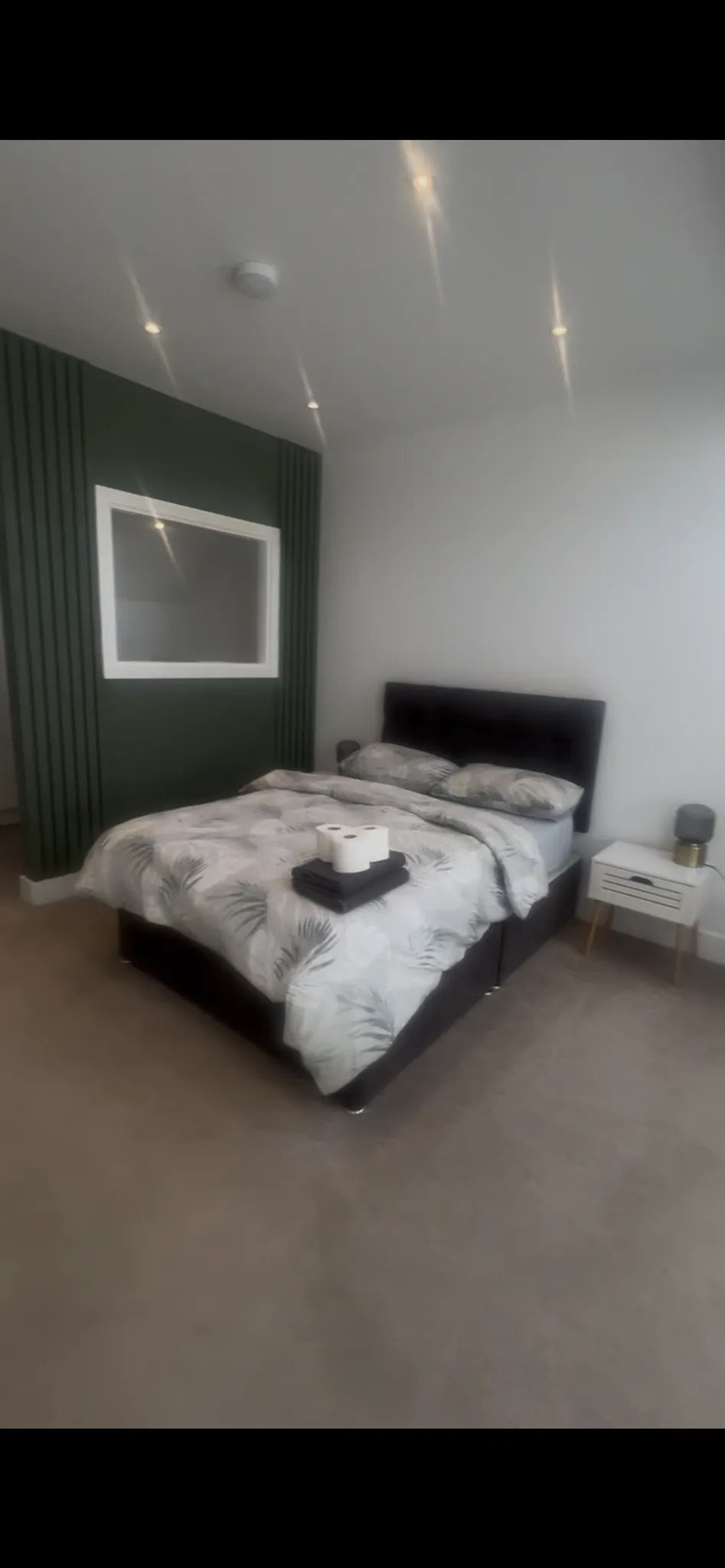 Bedroom with a double bed with patterned duvet, two pillows, a bedside table with a lamp, a green accent wall with an interior window, recessed ceiling lights and a smoke detector, and carpeted floor.