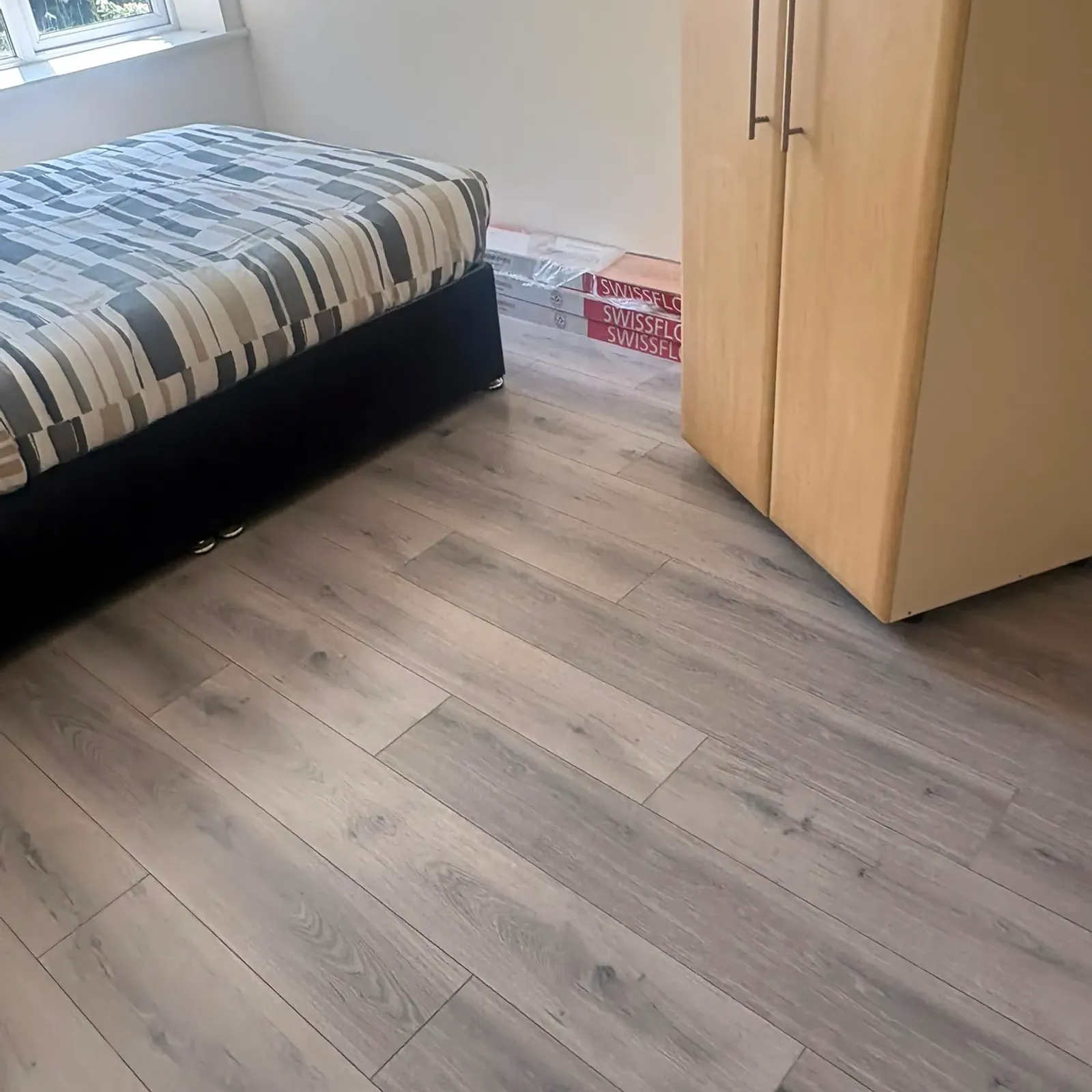 Bedroom with a double bed near a window, a wooden two-door wardrobe, and grey laminate flooring.