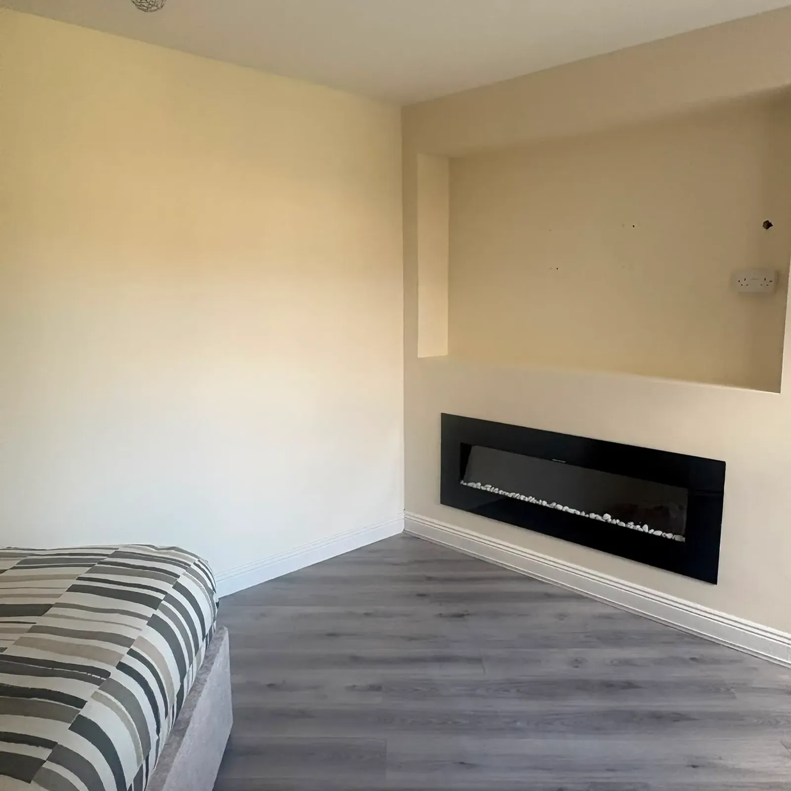 Bedroom corner with part of a striped bed visible, wall-mounted electric fireplace, pale walls and gray wood-look floor.