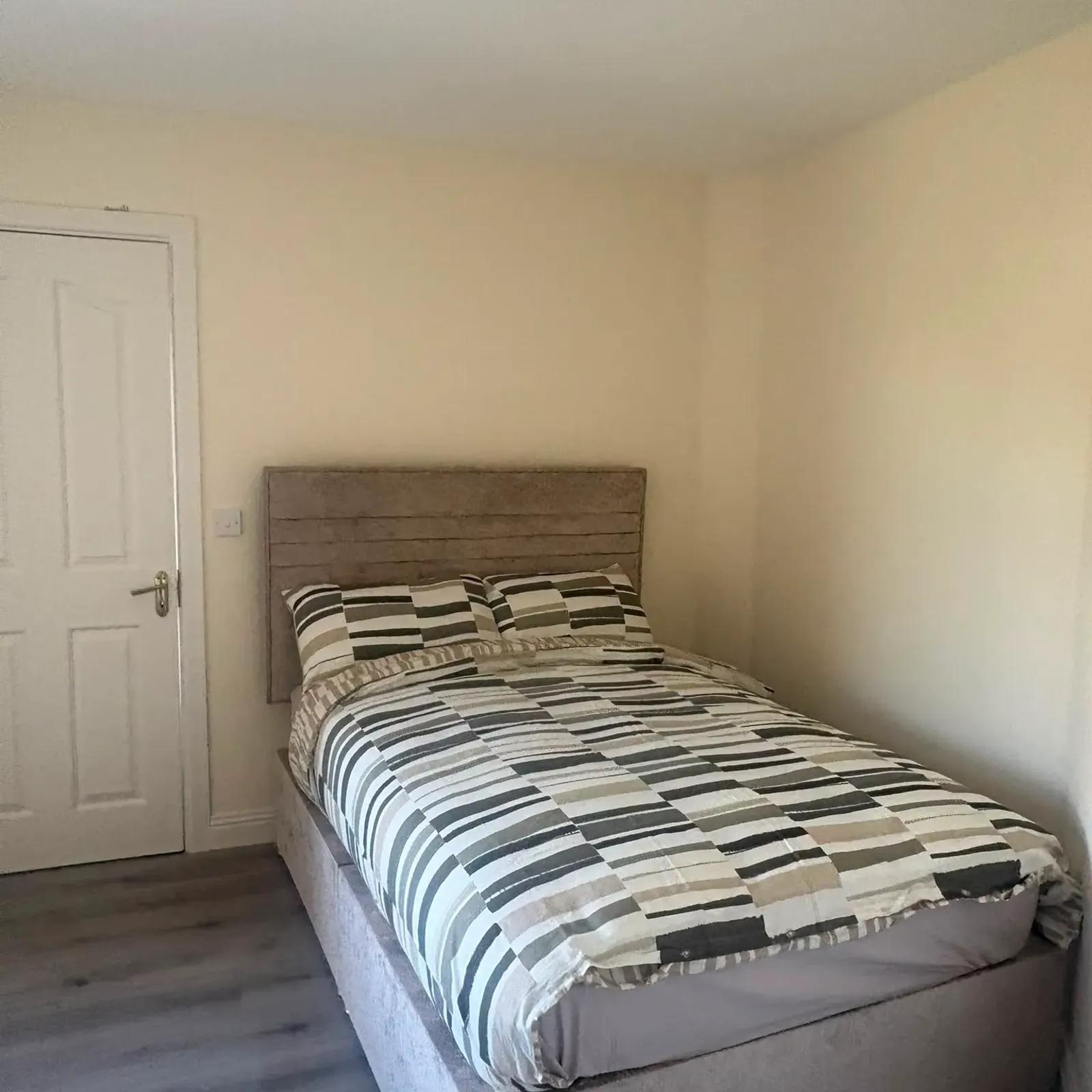 Bedroom with a single bed pushed against a corner, striped bedding, upholstered headboard, closed door and ceiling light fixture.