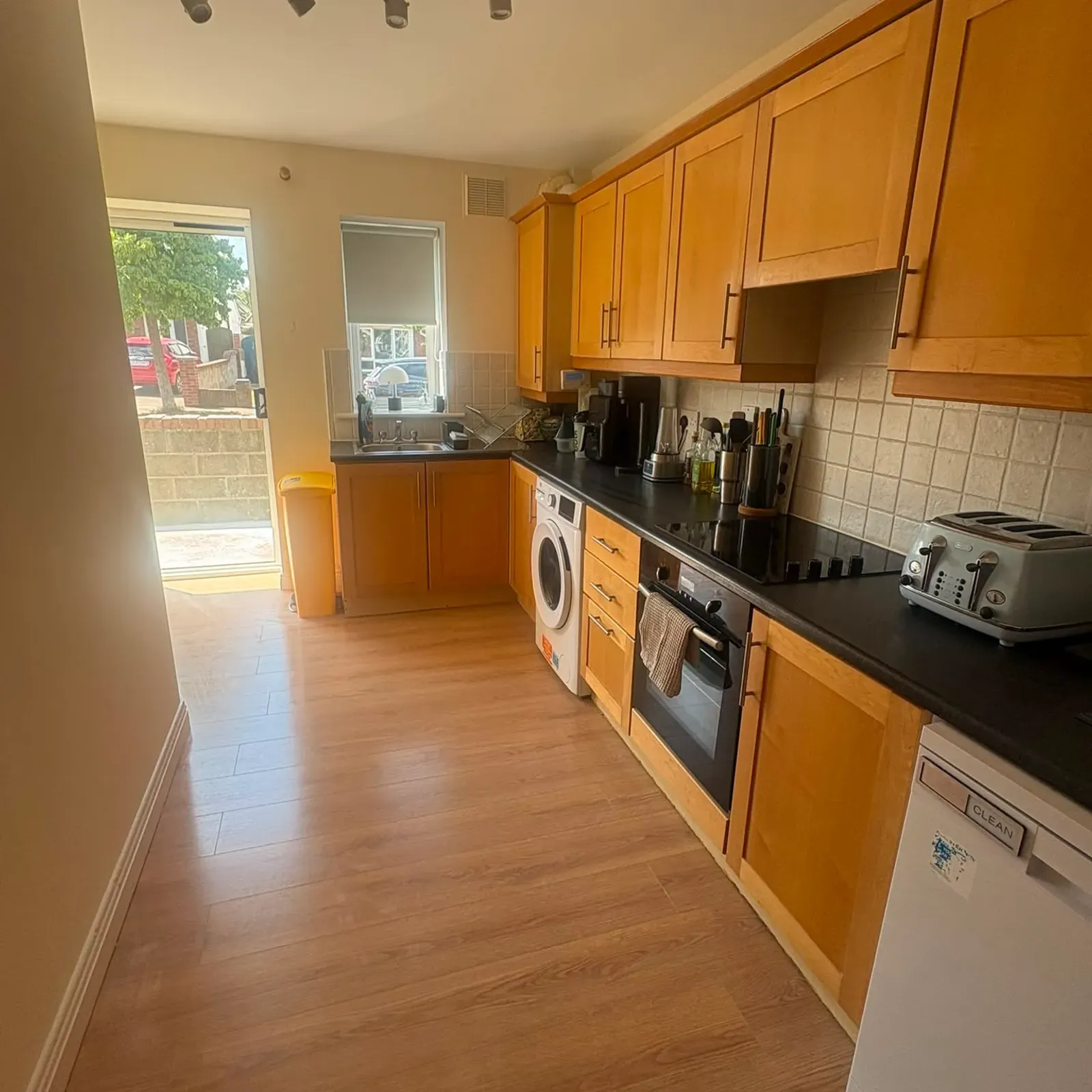 Kitchen with wooden cabinets, black countertop, sink under a small window, built-in oven, washing machine, toaster and a door to the outside.