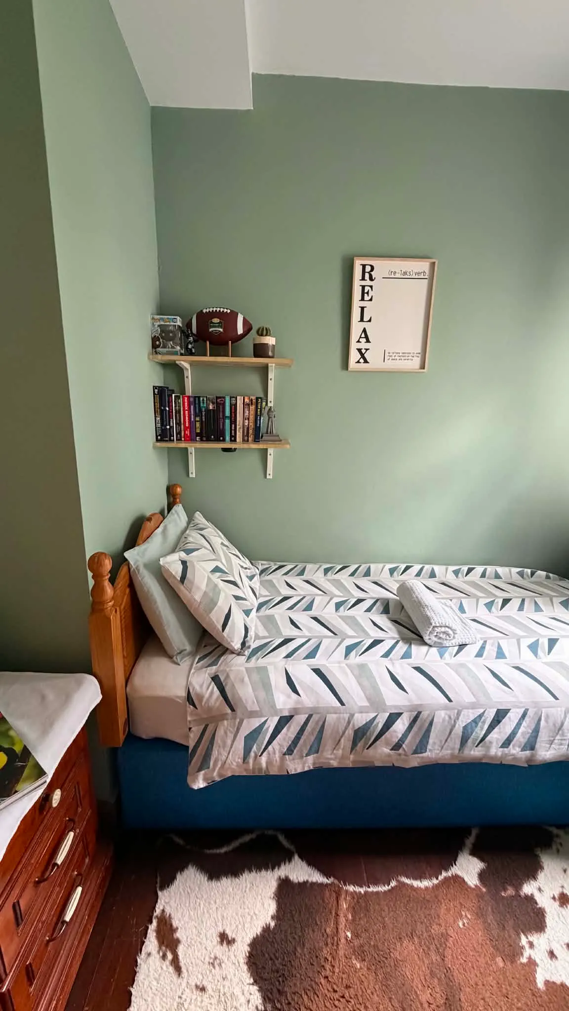 Bedroom with a single bed against a green wall, patterned bedding, pillows and a folded towel; floating shelves with books and a football and framed wall art reading 'RELAX'.