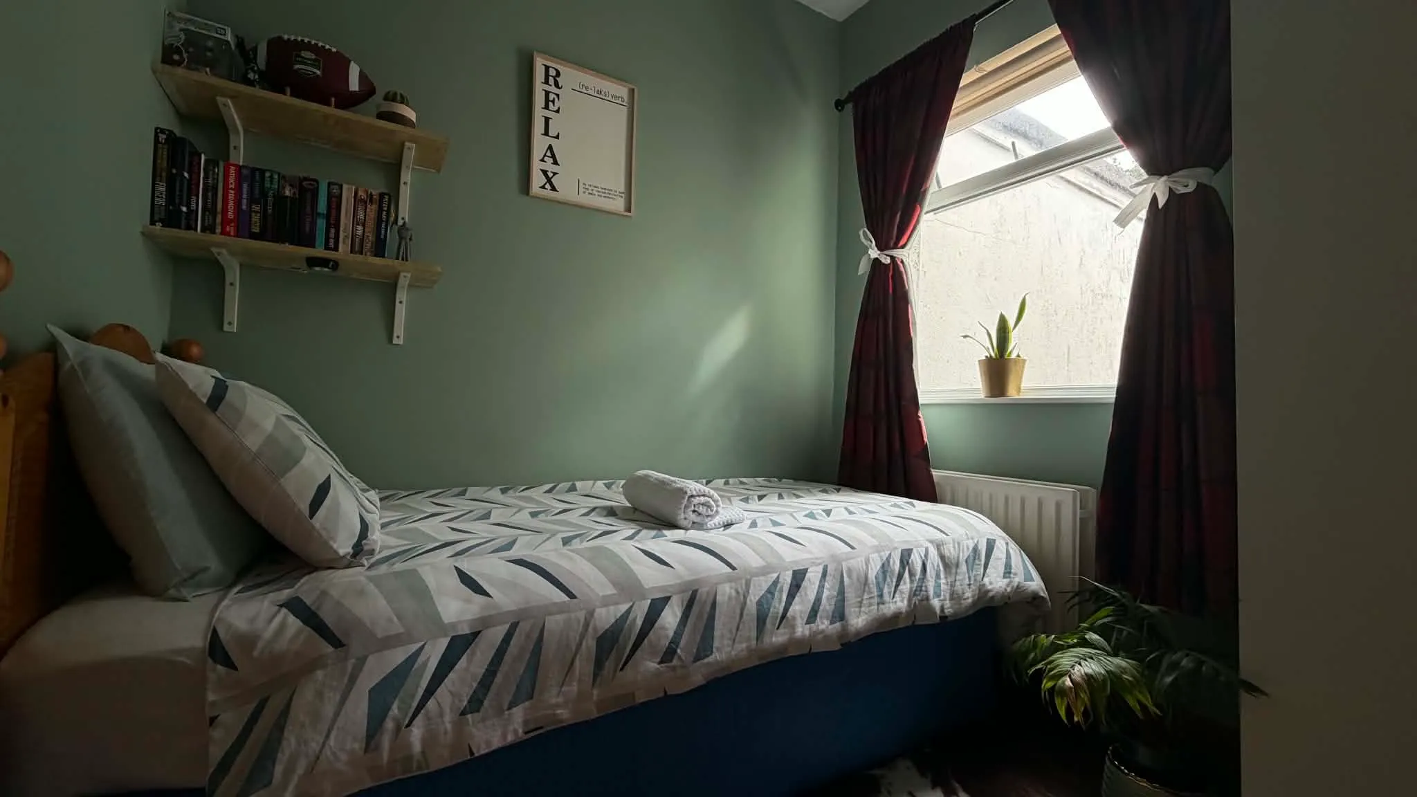 Bedroom with a single bed and patterned bedding, pillows, wall shelves with books, a framed 'RELAX' print, a window with dark curtains and a small potted plant on the sill.