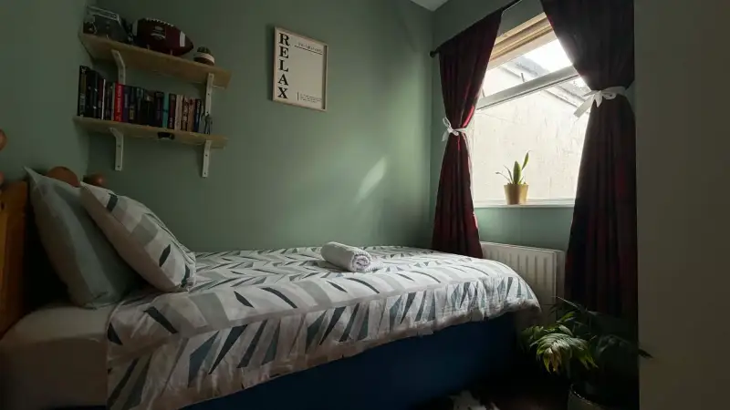 Bedroom with a single bed and patterned bedding, pillows, wall shelves with books, a framed 'RELAX' print, a window with dark curtains and a small potted plant on the sill.