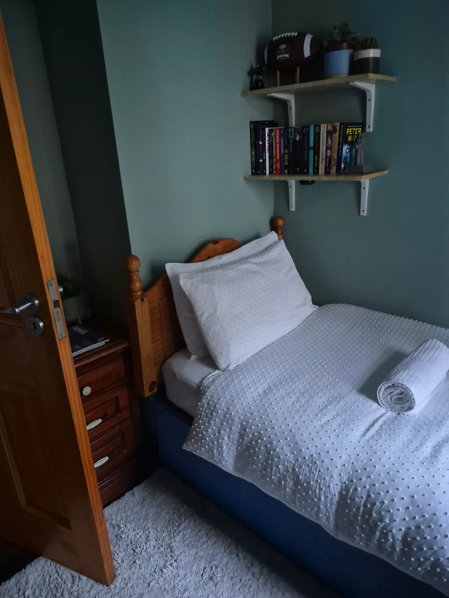 Bedroom with a single bed, white pillow and textured white bedding, wooden headboard, two wall-mounted shelves with books and small items, and an open wooden door.