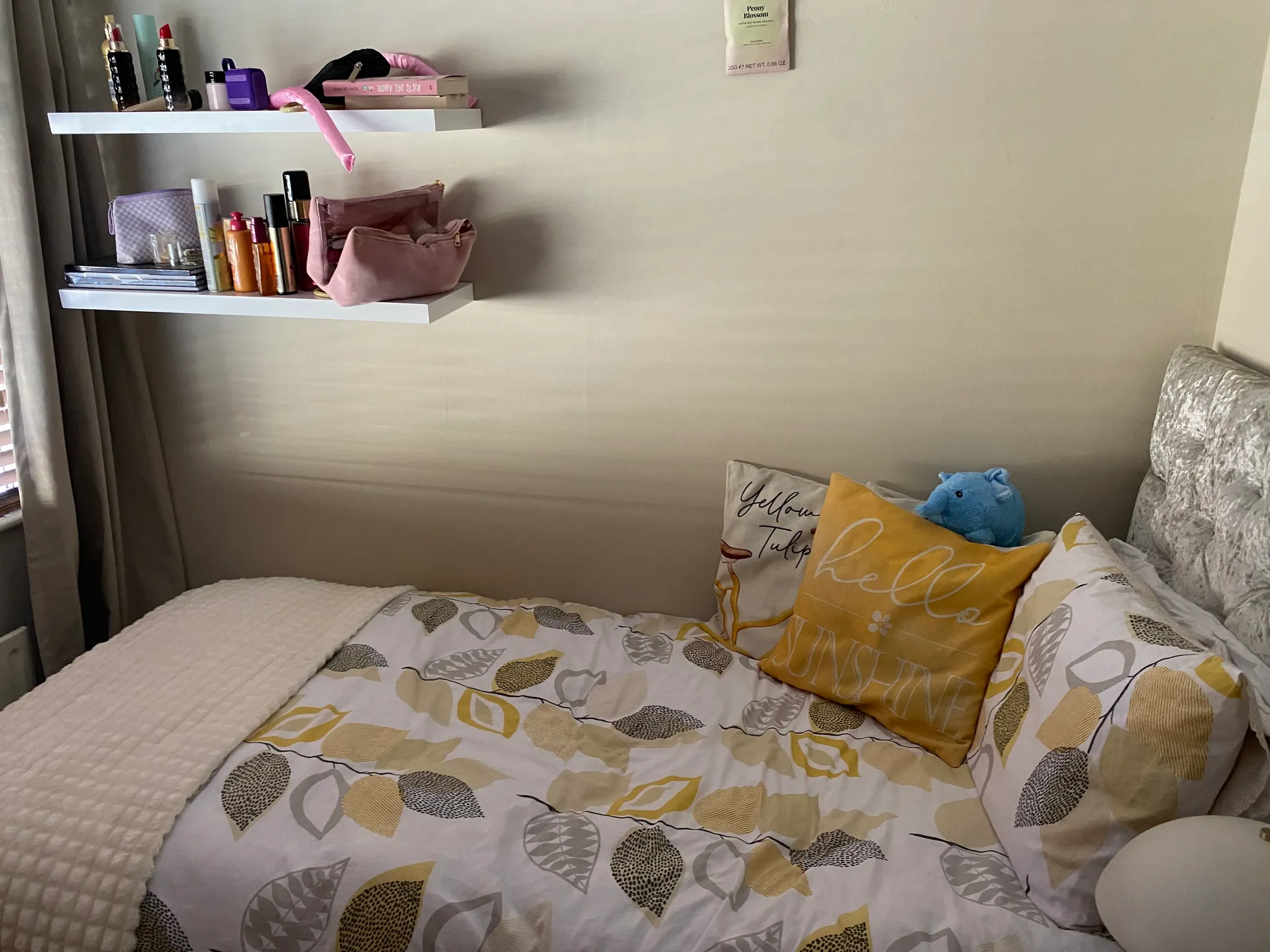 Bedroom corner with a bed, patterned duvet and several pillows, wall-mounted shelves holding personal items and cosmetics, and curtains at the window.