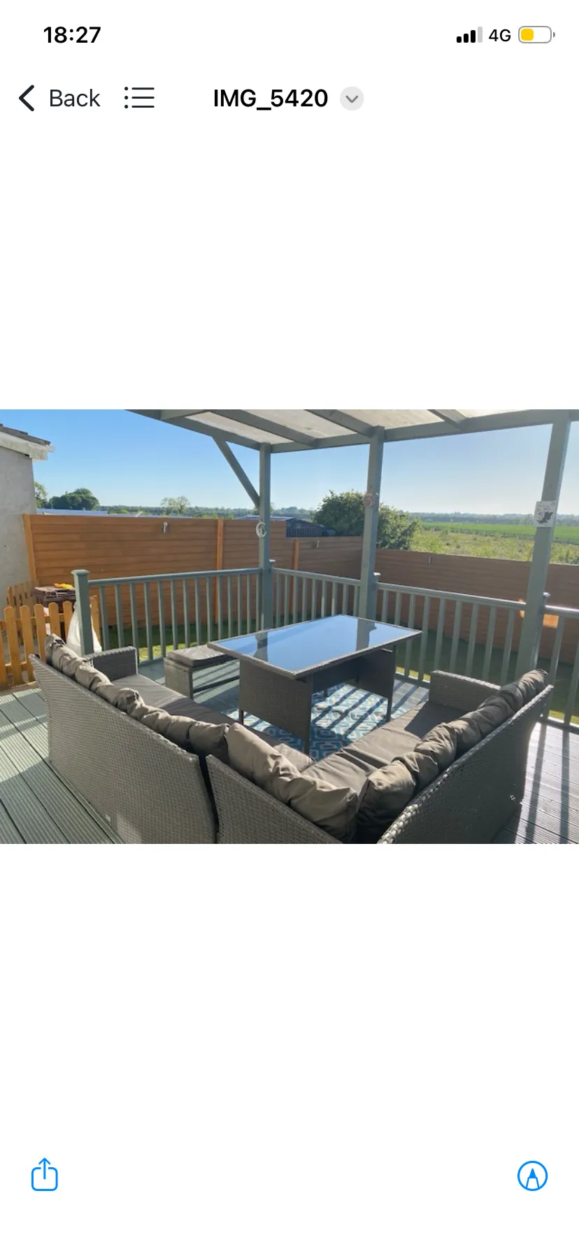 Covered outdoor deck with a woven sectional sofa facing a glass-top coffee table, wooden railing and fence, and a view of fields beyond.
