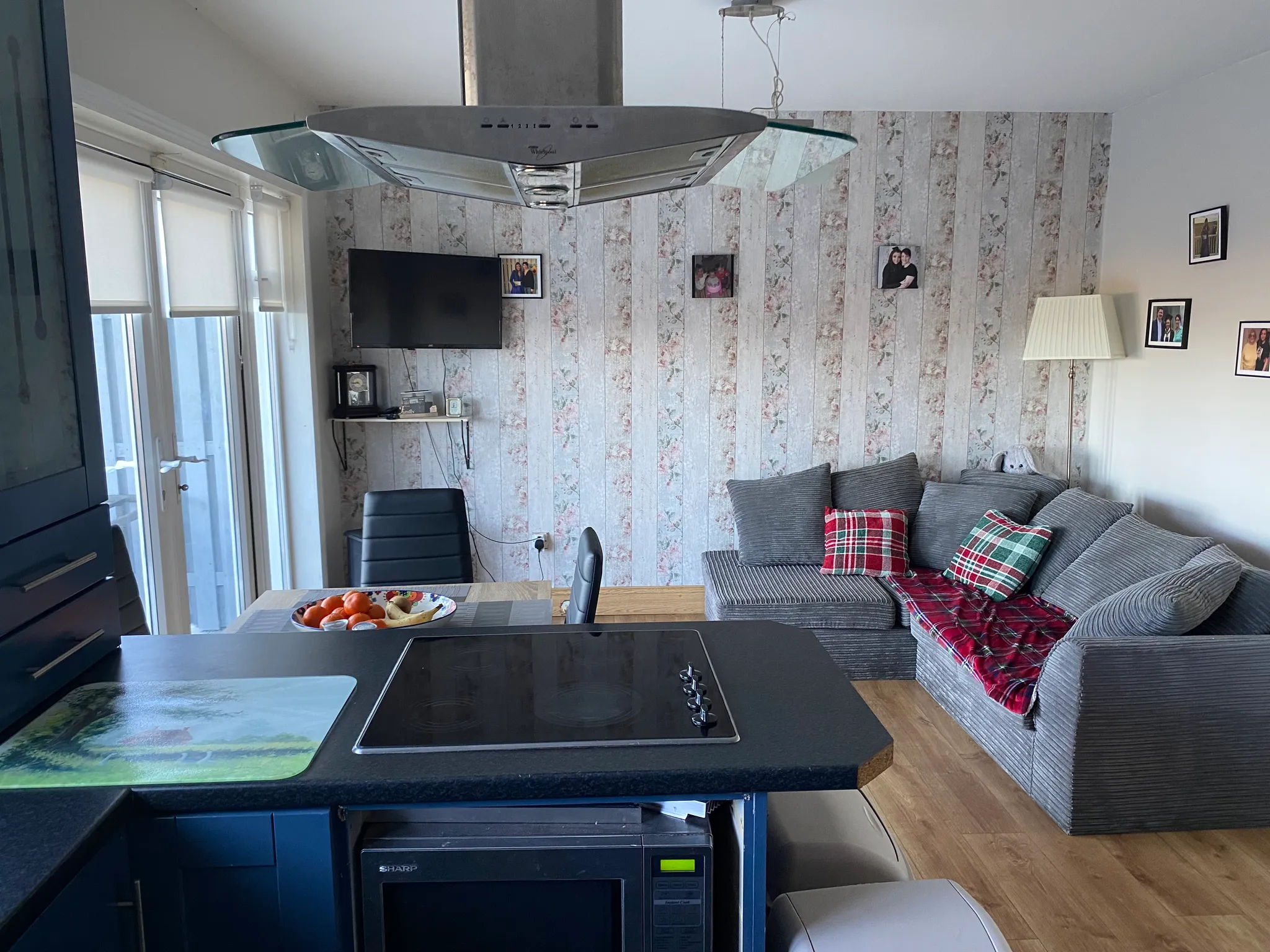 Open-plan living area with a grey upholstered corner sofa with patterned cushions, a floor lamp, wall-mounted TV, dining table with fruit bowl, and a kitchen island with an induction hob and overhead extractor hood.