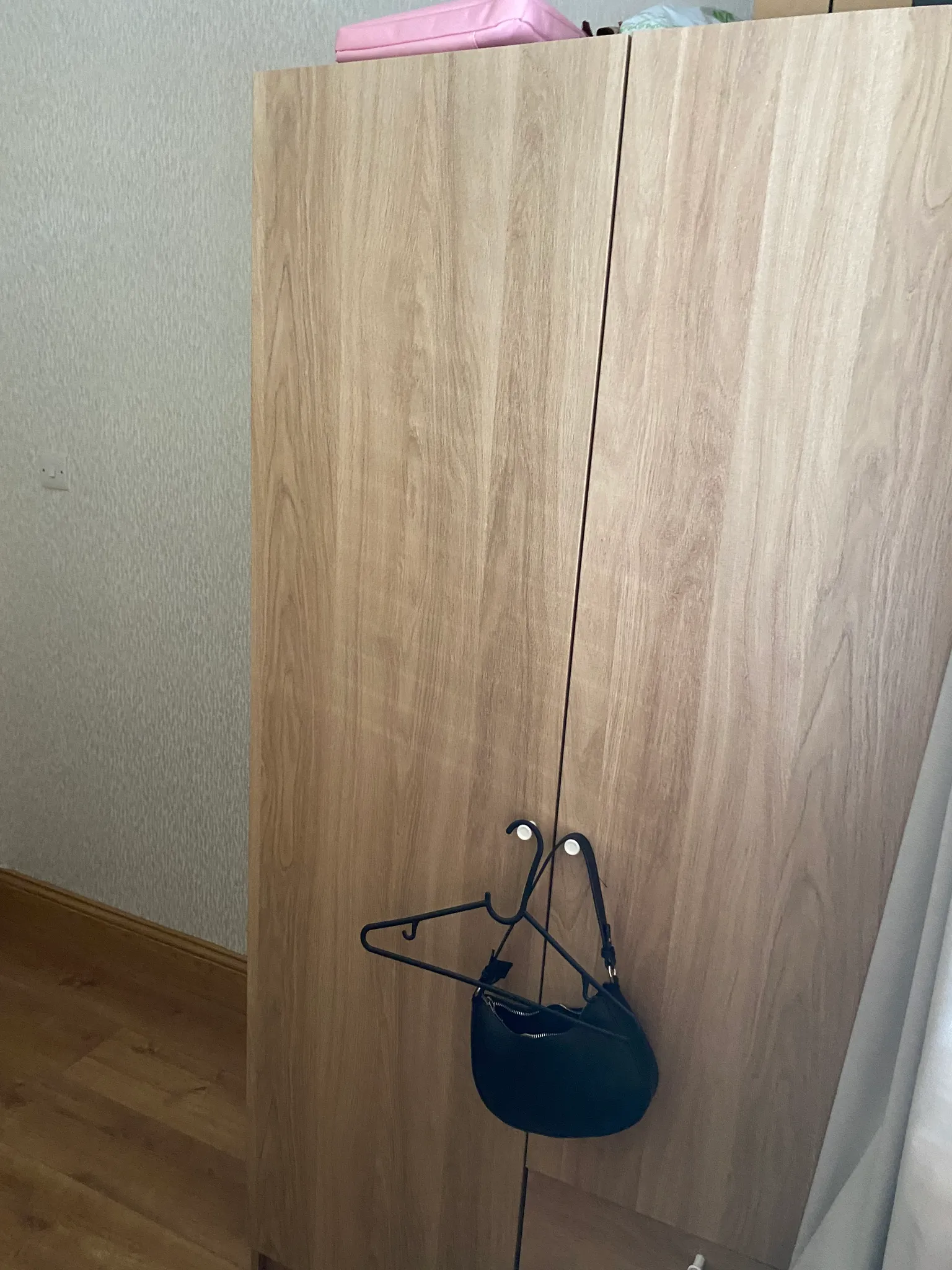 Partial view of a wooden wardrobe with two doors, hangers and a small black bag hanging from a hook, adjacent wall and floor visible.