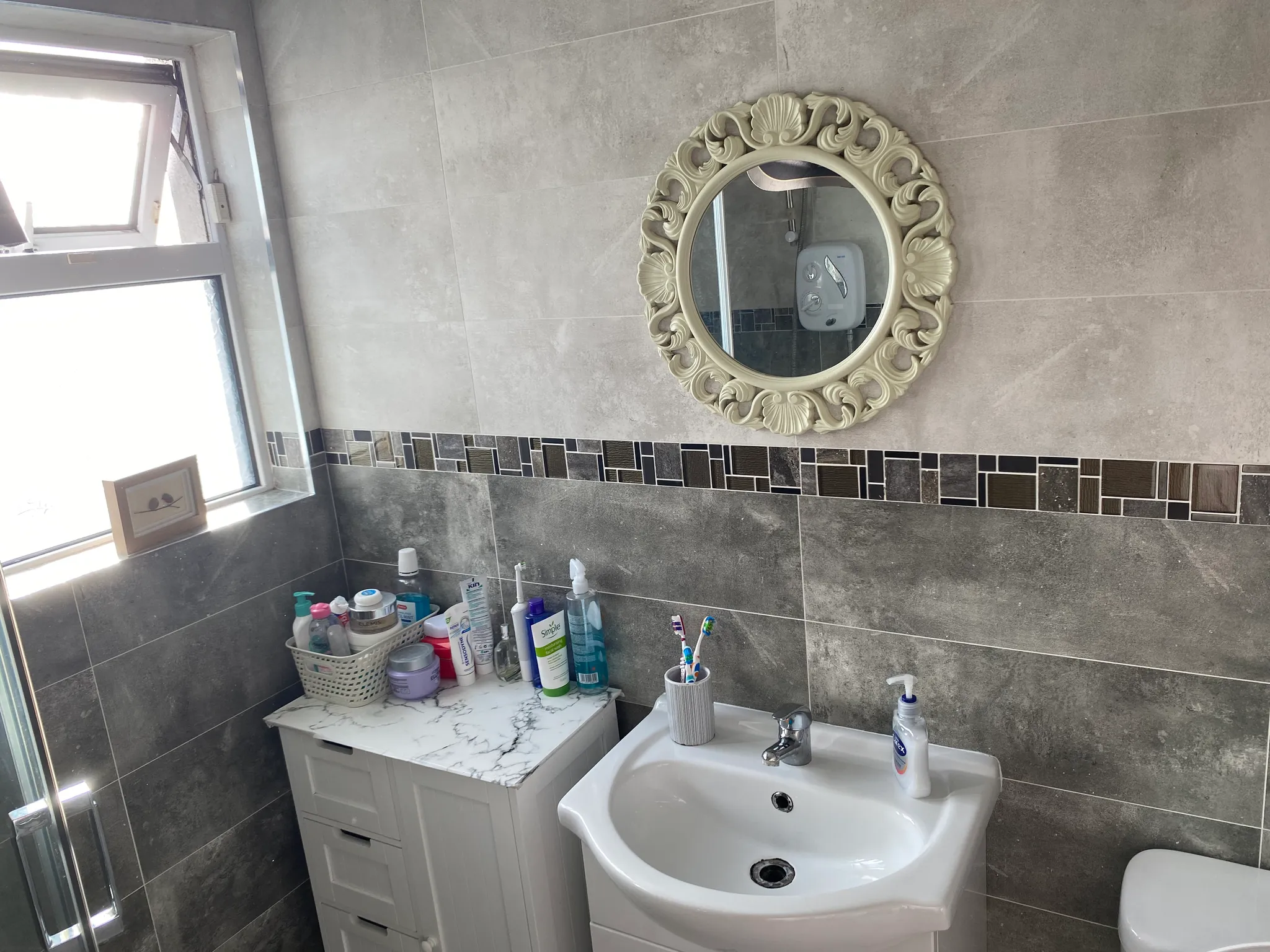 Bathroom with a sink, ornate round mirror on a tiled wall, a small storage cabinet and toiletries on the countertop near a window.