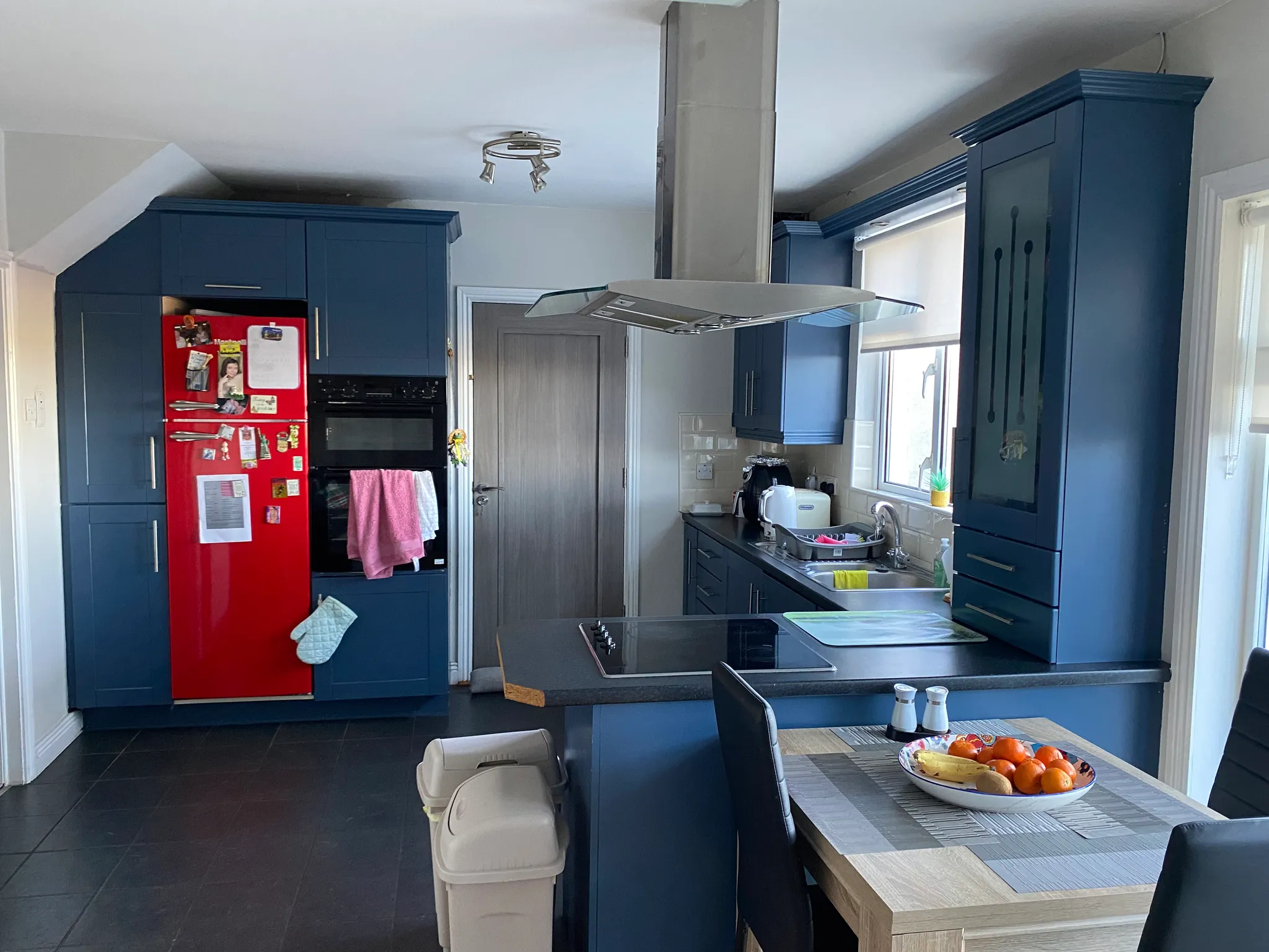 Kitchen with blue cabinets, red refrigerator, island cooktop with stainless steel extractor hood, sink under a window and a dining table with a bowl of fruit.