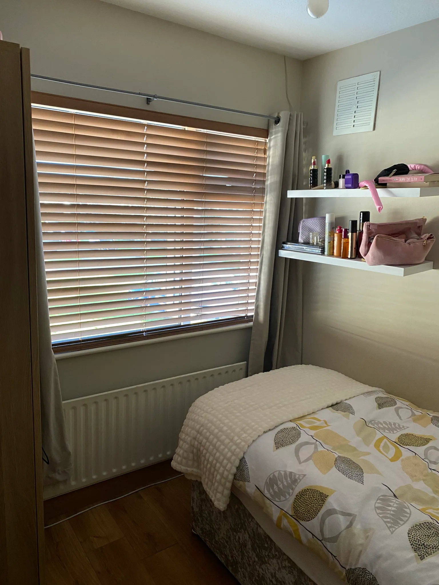 Bedroom with a bed in the foreground, a large window with closed wooden blinds and a curtain, and two wall shelves holding toiletry items and small bags.