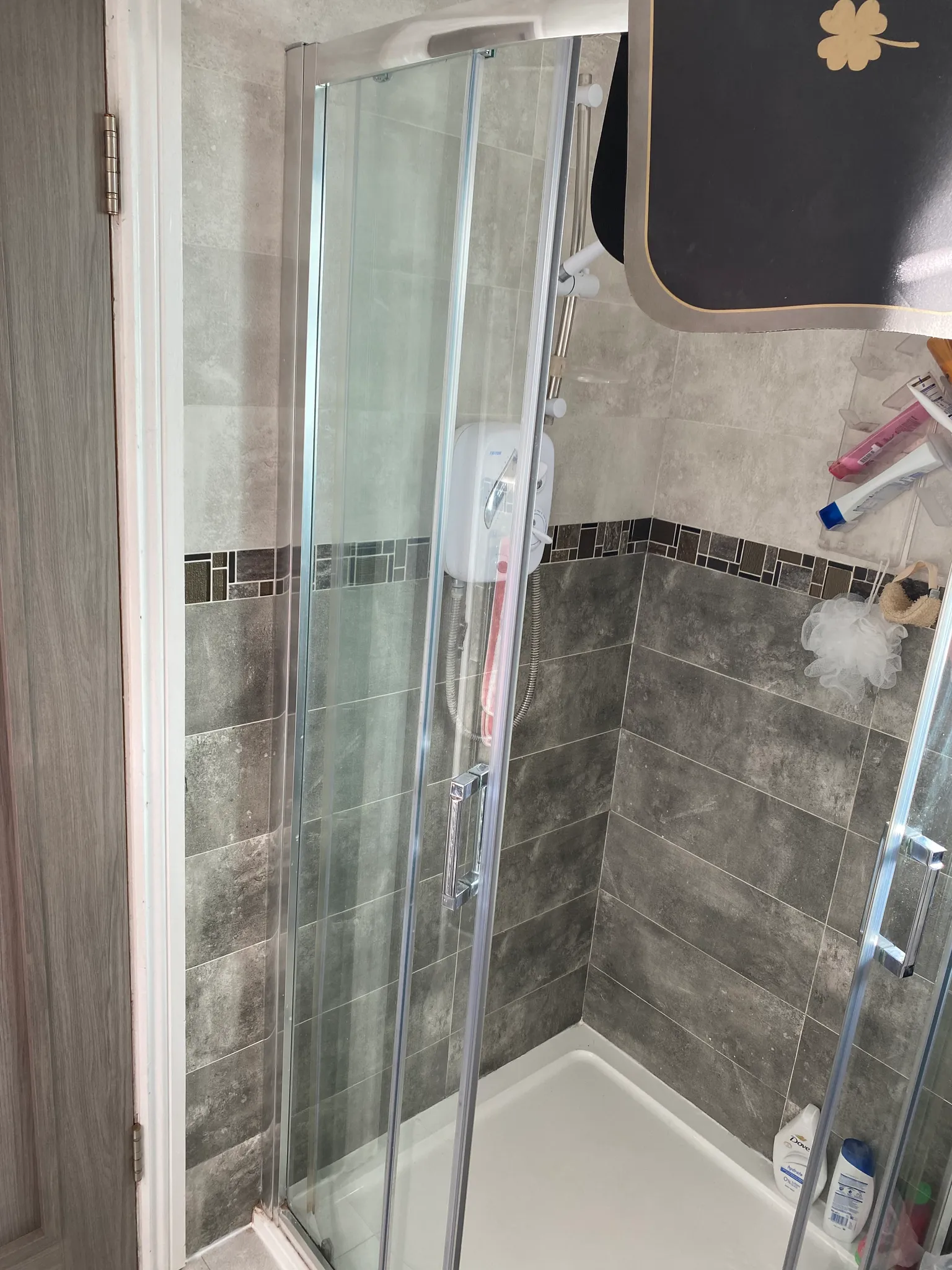 Bathroom shower stall with glass sliding doors, tiled walls, wall-mounted electric shower unit and toiletries on a corner shelf.