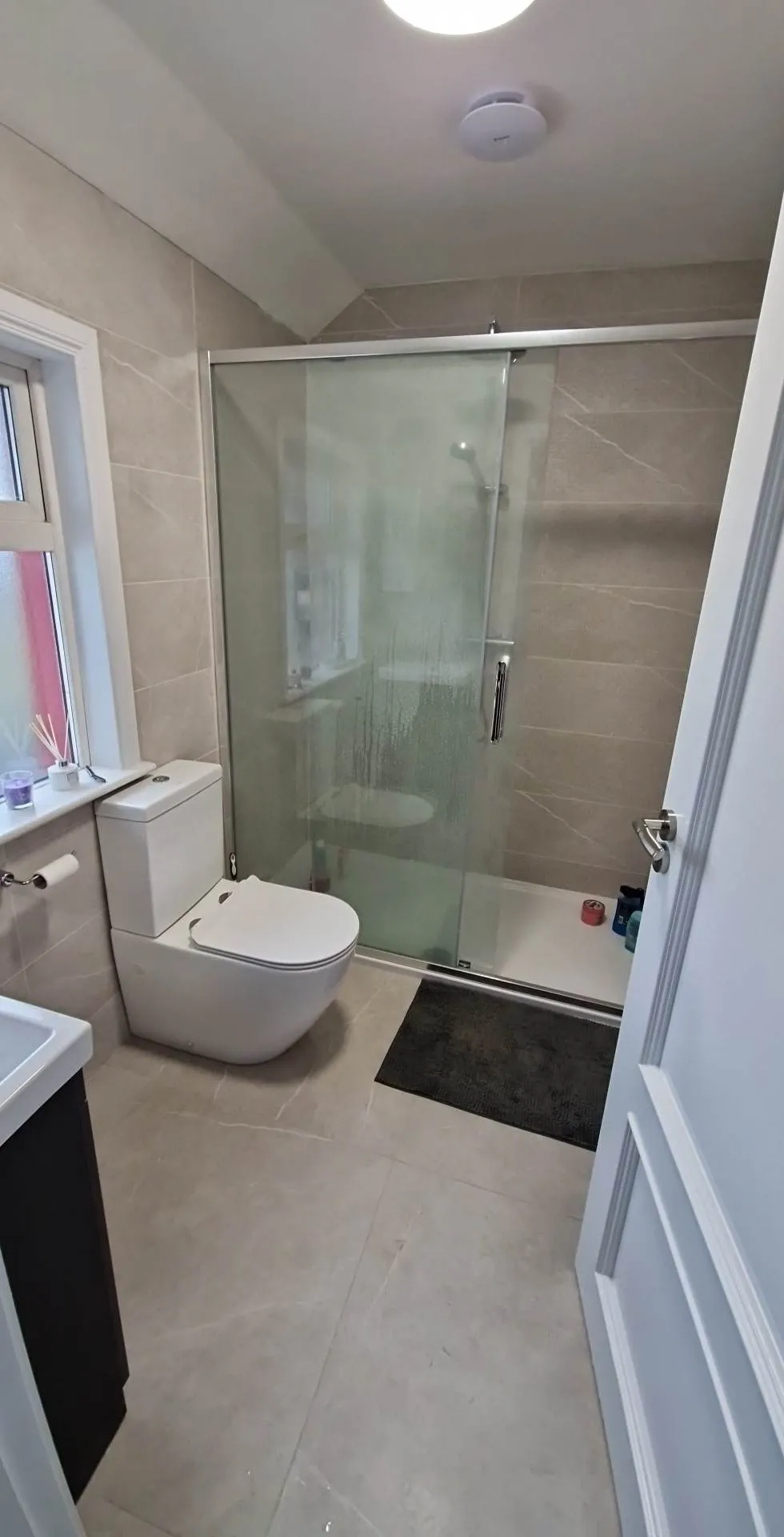 Bathroom with a toilet next to a frosted glass shower enclosure, window on the left, and a bath mat on the floor