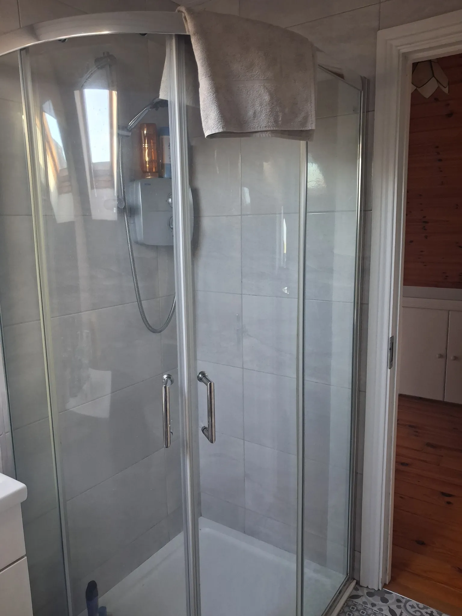 Bathroom with a corner glass shower enclosure, handheld shower unit, and a towel draped over the top of the doors.
