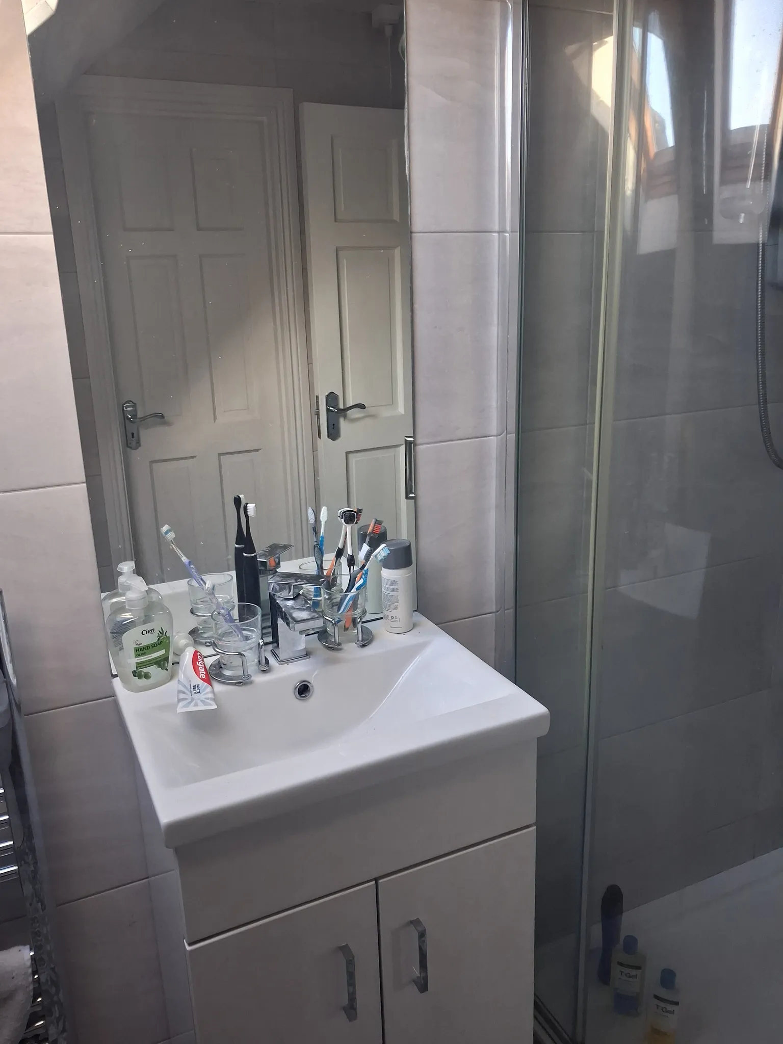 Bathroom sink and mirror with toothbrushes and toiletries on the vanity, tiled wall and glass shower door to the right.