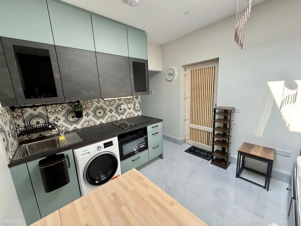 Kitchen with mint and dark cabinets, patterned tiled backsplash, sink, washing machine, oven, and countertop with wooden table in foreground.