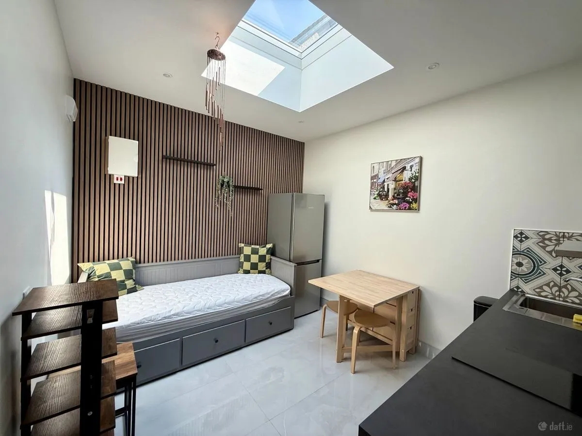 Room with a single daybed with storage drawers, skylight, wooden slatted accent wall, small dining table with two chairs, and a kitchenette area with refrigerator.