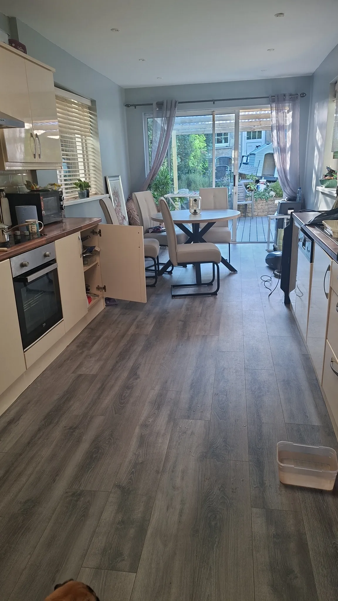 Open-plan kitchen and dining area with a round table and four chairs, cabinets and oven on the left, sliding glass door to a garden at the rear.