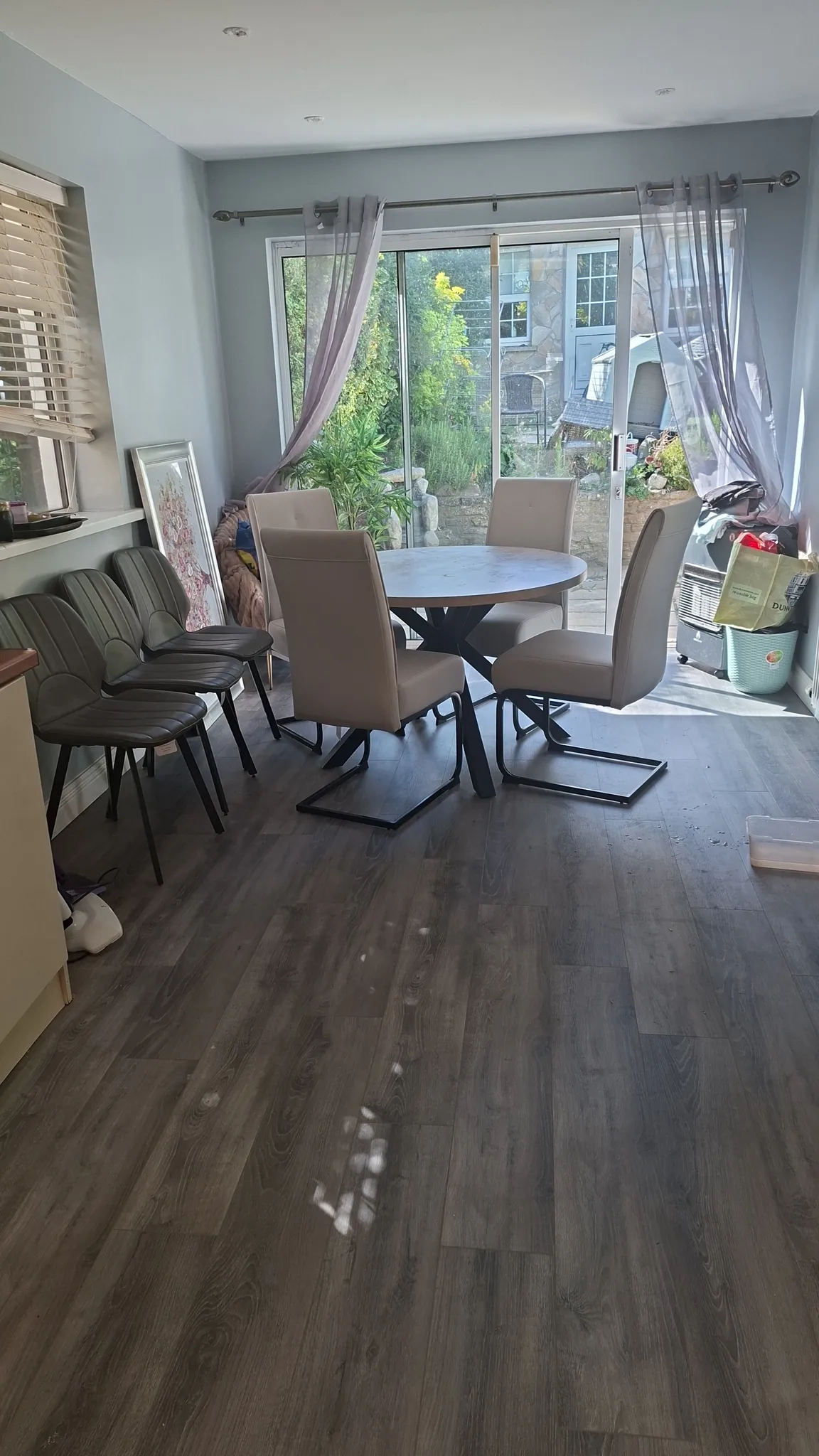 Dining room with a round table and four chairs near sliding glass doors with sheer curtains, additional stacked chairs against the left wall and items stored near the right corner.