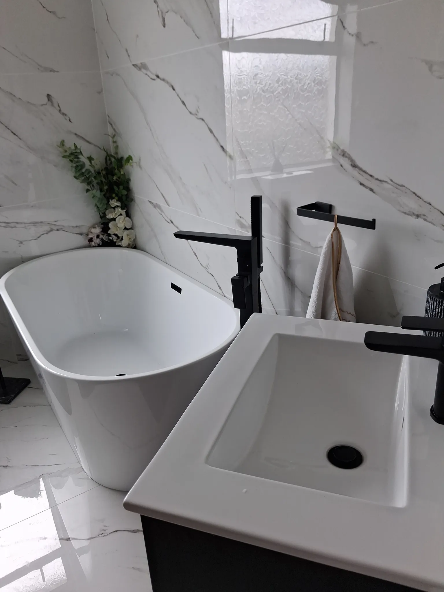 Bathroom with a freestanding bathtub, wall-mounted black faucet, sink and towel on a wall hook, and marble-look tiled walls with a small frosted window.