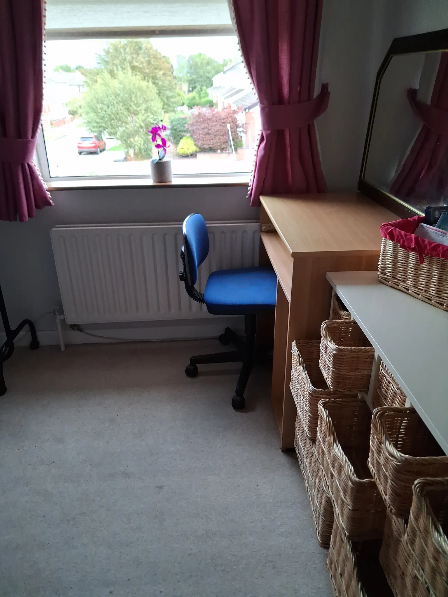 Small workspace with a wooden desk, blue swivel chair, window with pink curtains and a small potted plant on the sill, mirror and stacked wicker storage baskets to the right.