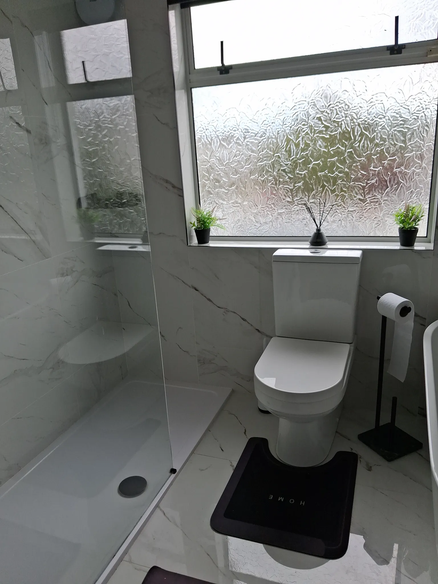 Bathroom with a toilet next to a frosted window, small potted plants on the sill, a glass shower partition and tiled walls and floor.