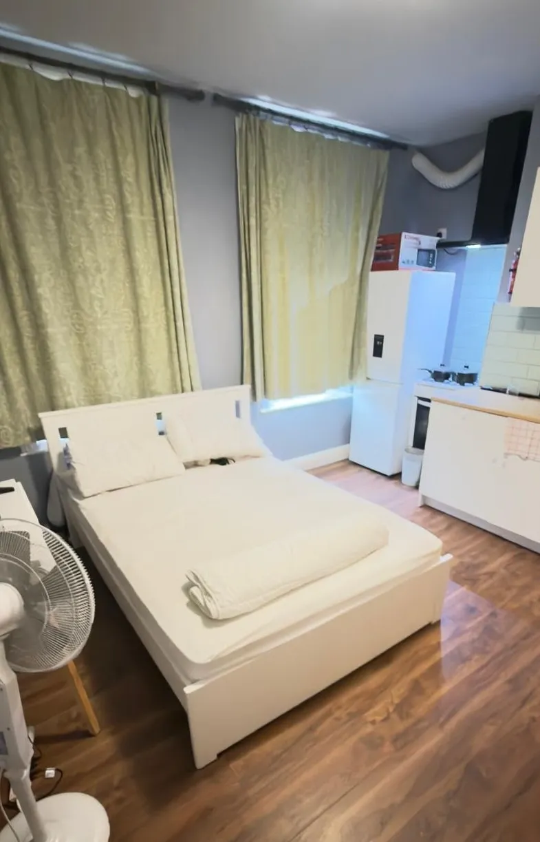 Bedroom with a double bed, pillows, folded blanket, window with drawn curtains, standing fan, and a small kitchenette with refrigerator in the corner.